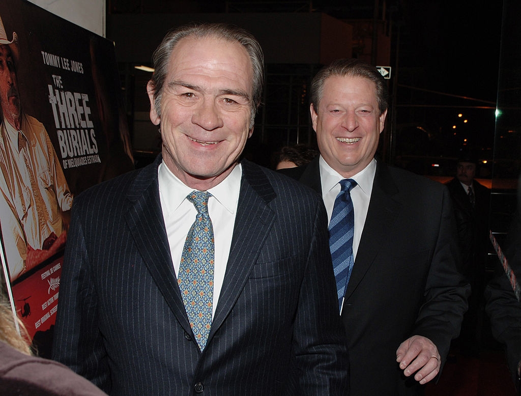 Two men in suits smile at an event. A poster is partially visible in the background