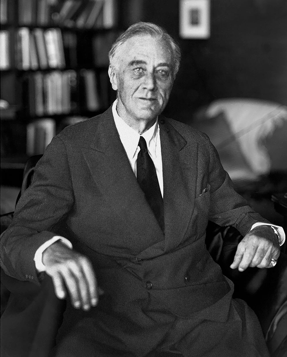 An older man in a suit and tie sits in a chair in a room with bookshelves and a framed picture