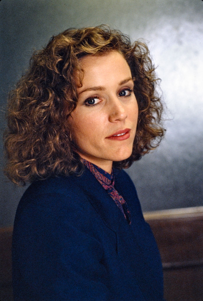 A person with curly hair is wearing a suit and patterned tie, looking towards the camera with a neutral expression