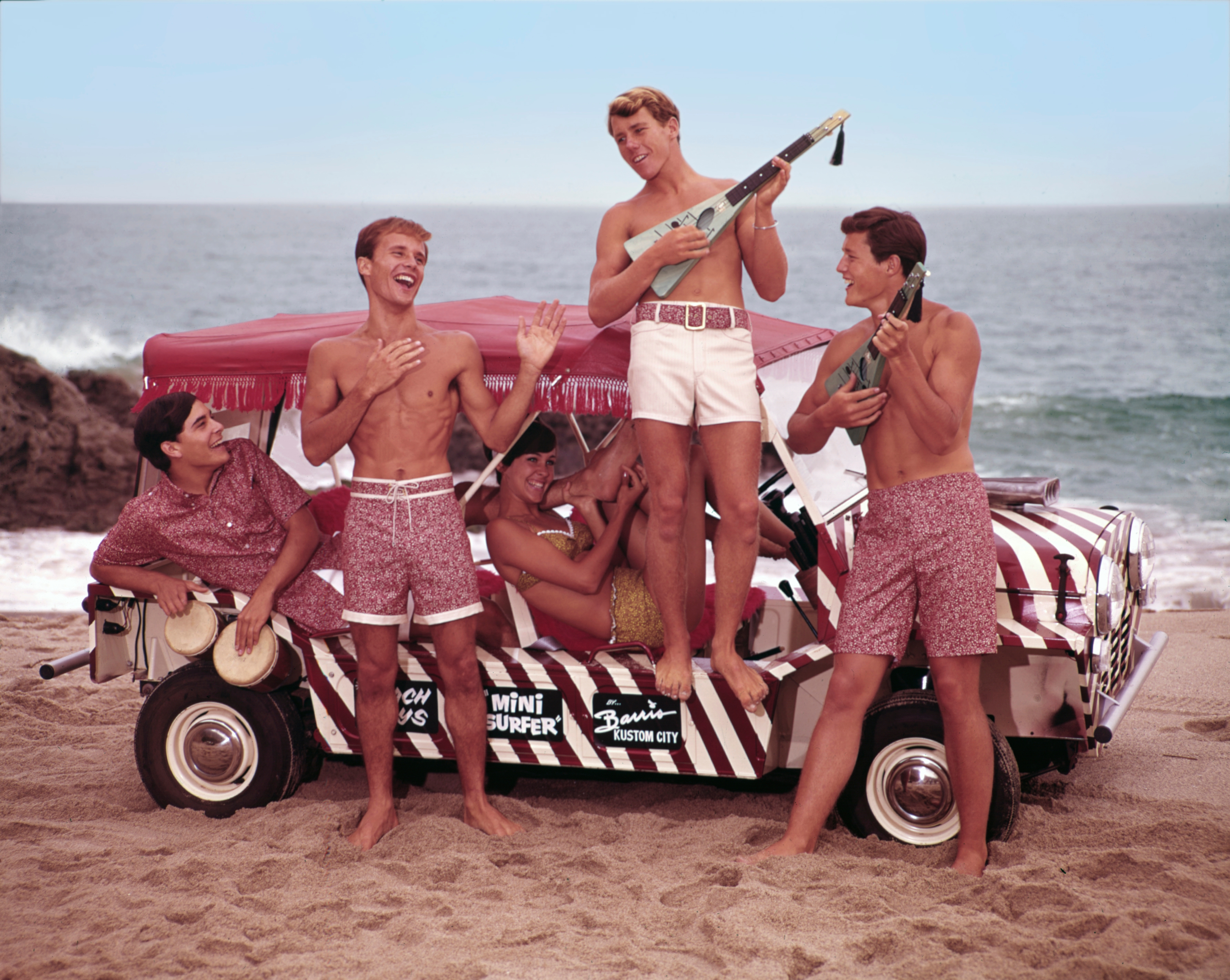 Five people in swimwear stand and sit on a decorated beach buggy, playing instruments and laughing by the ocean