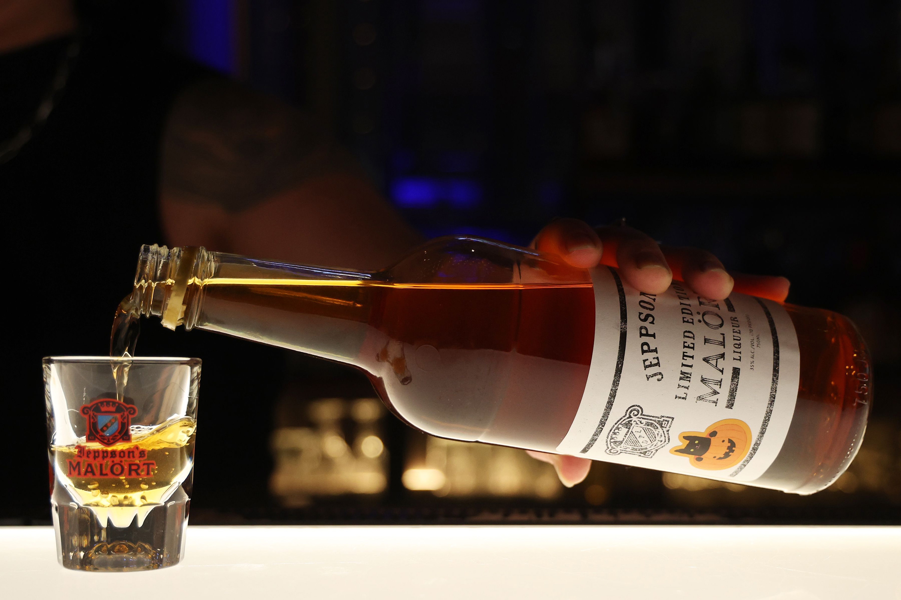 A hand pours Jeppson's Malört into a branded shot glass on a well-lit bar counter