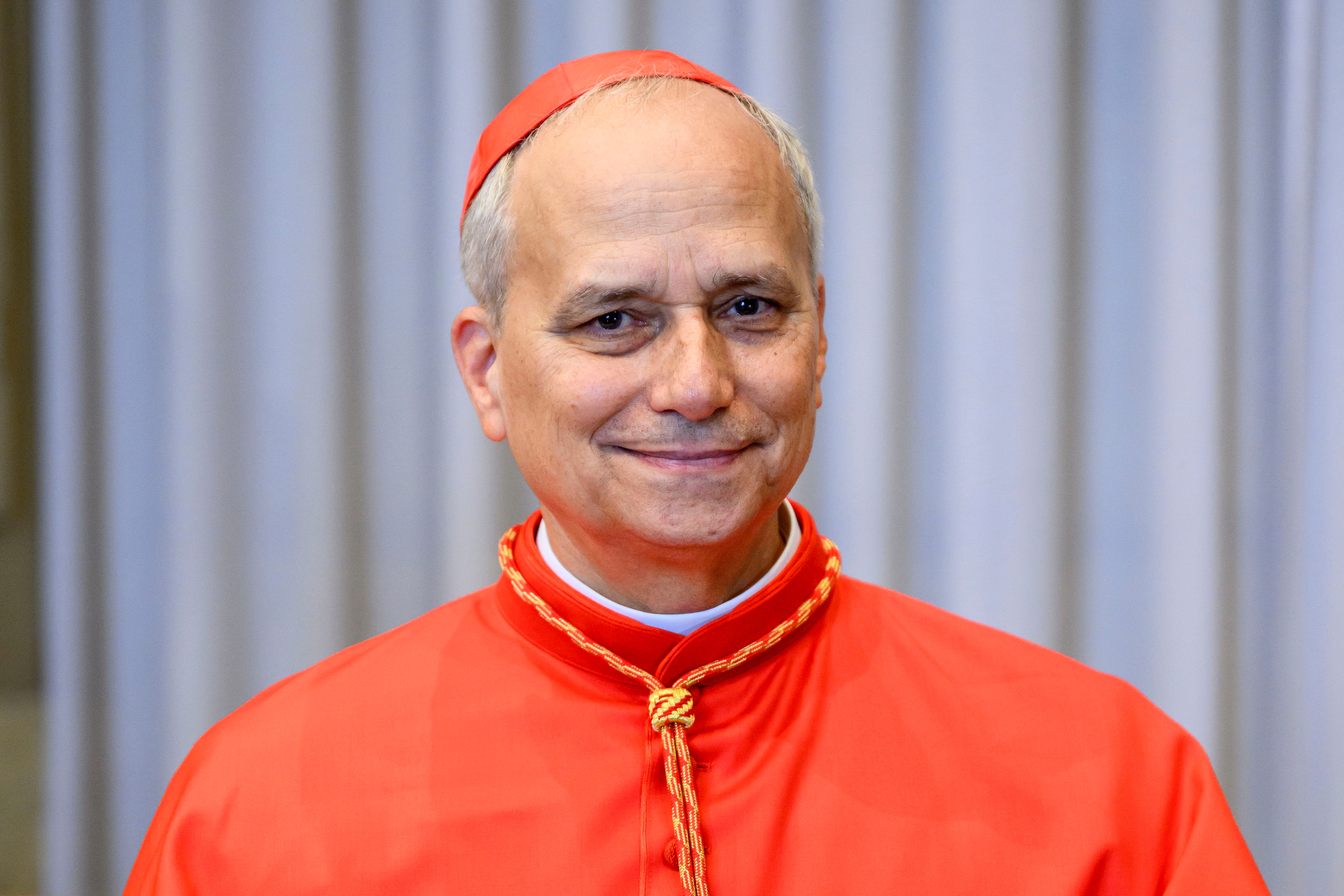 A man wearing a clerical robe and cap stands and smiles, suggesting a formal or religious context