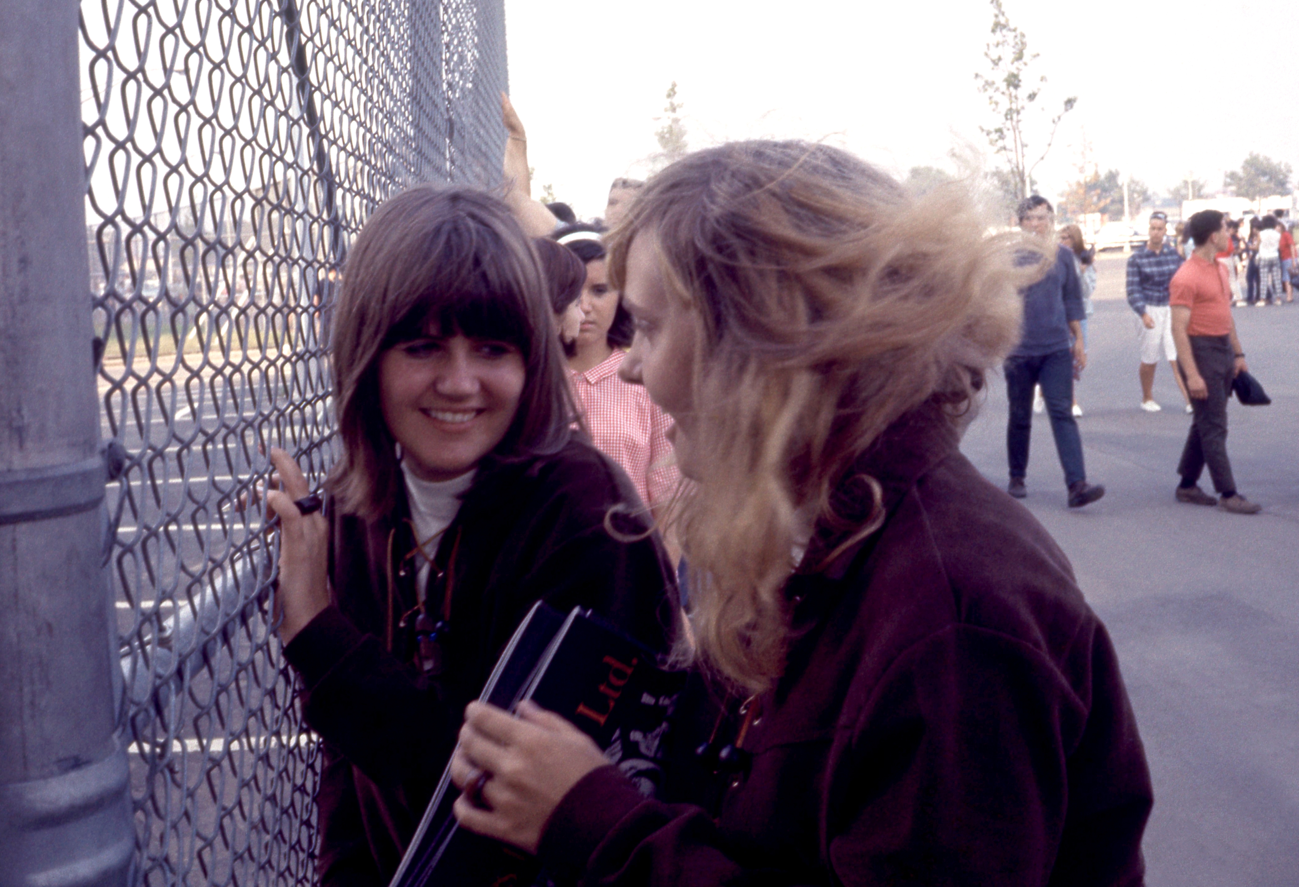 Two women converse by a chain-link fence. One smiles while the other gestures, holding a book. People walk in the background