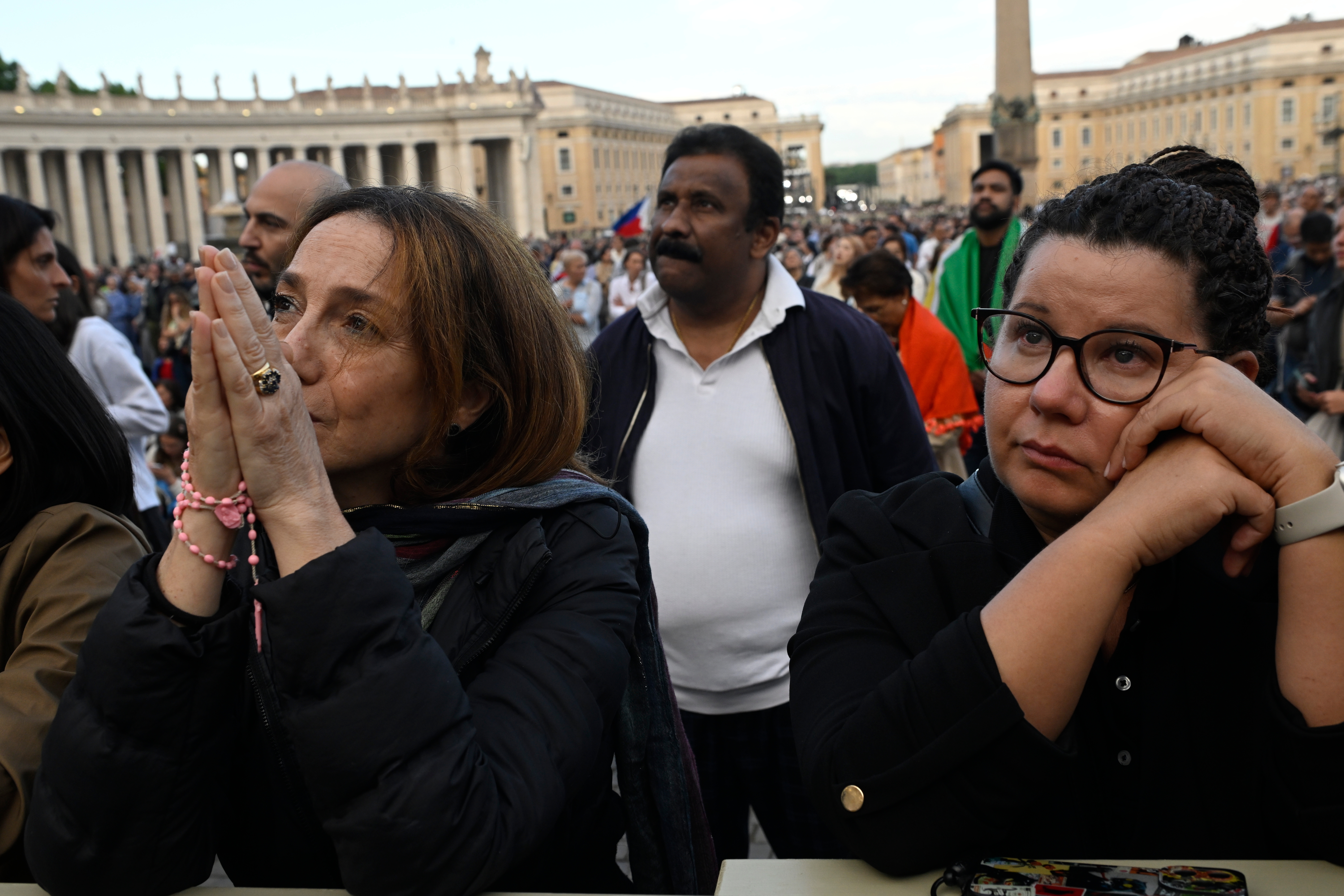 A crowd gathered in a public square, with people showing expressions of anticipation and concern. A woman holds rosary beads in her hands