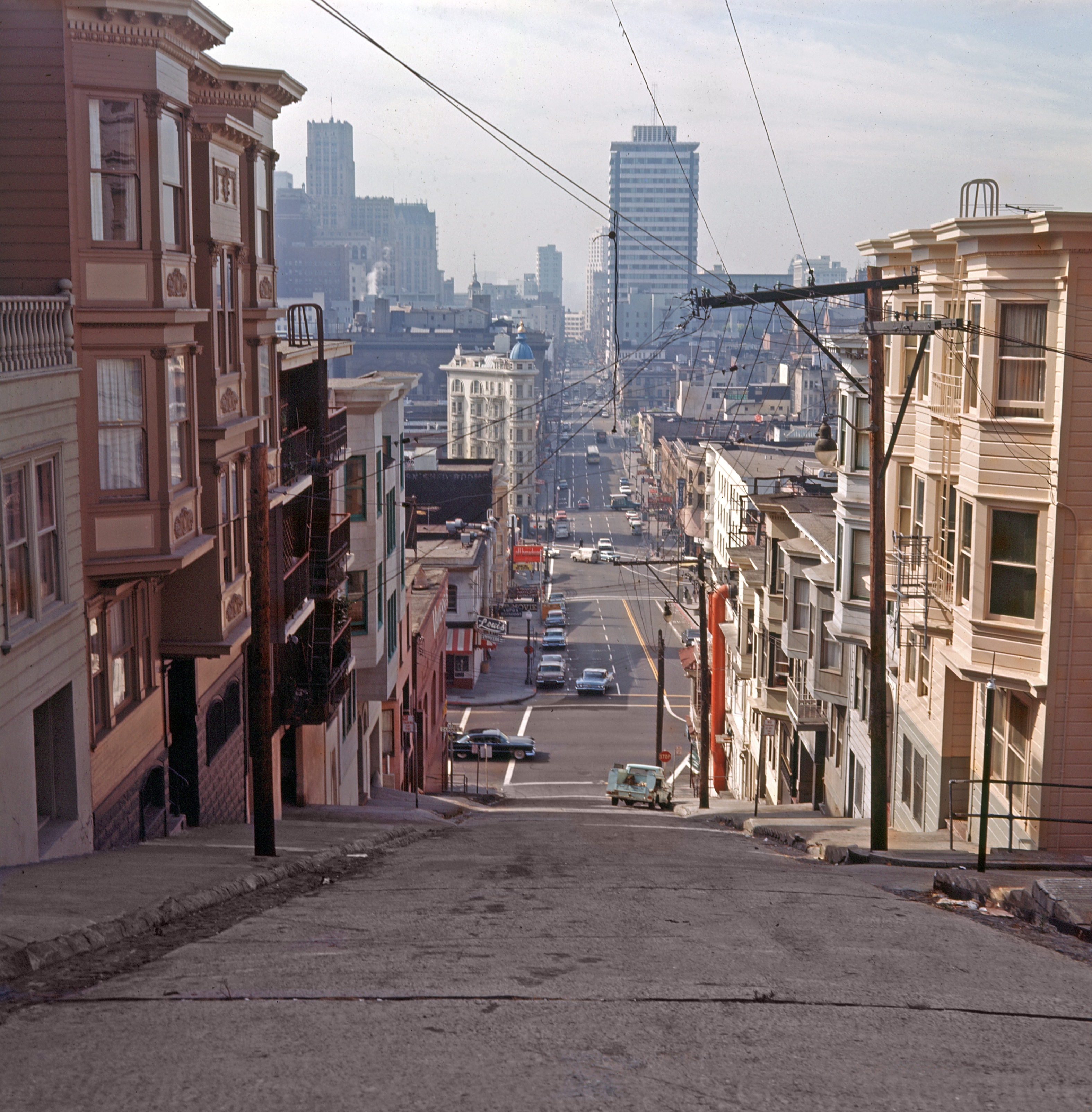 Steep urban street view with cars and buildings, leading to a distant city skyline under a clear sky