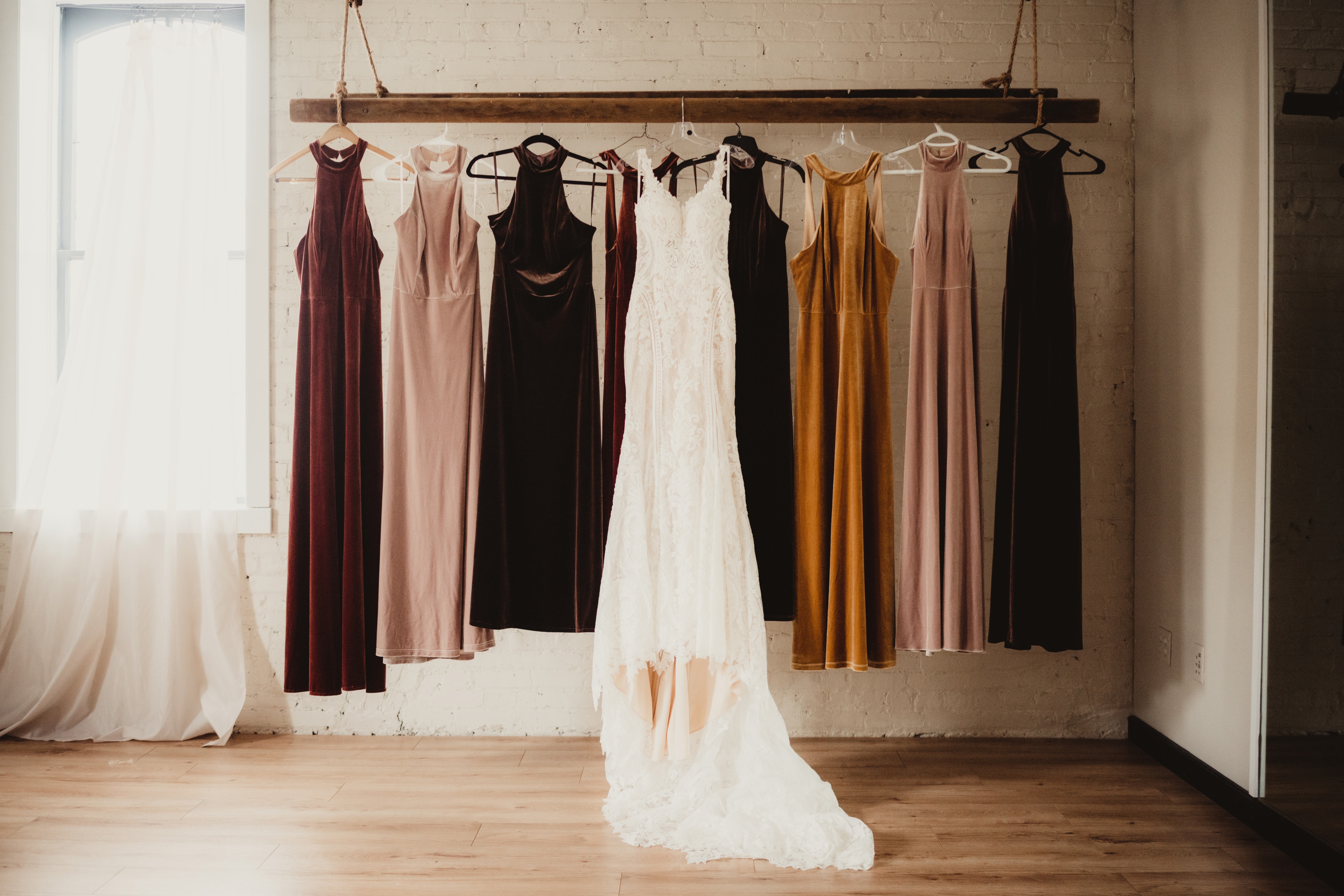 A row of elegant velvet gowns on hangers, with a white lace wedding dress centered, hanging from a wooden rack in a fashion display