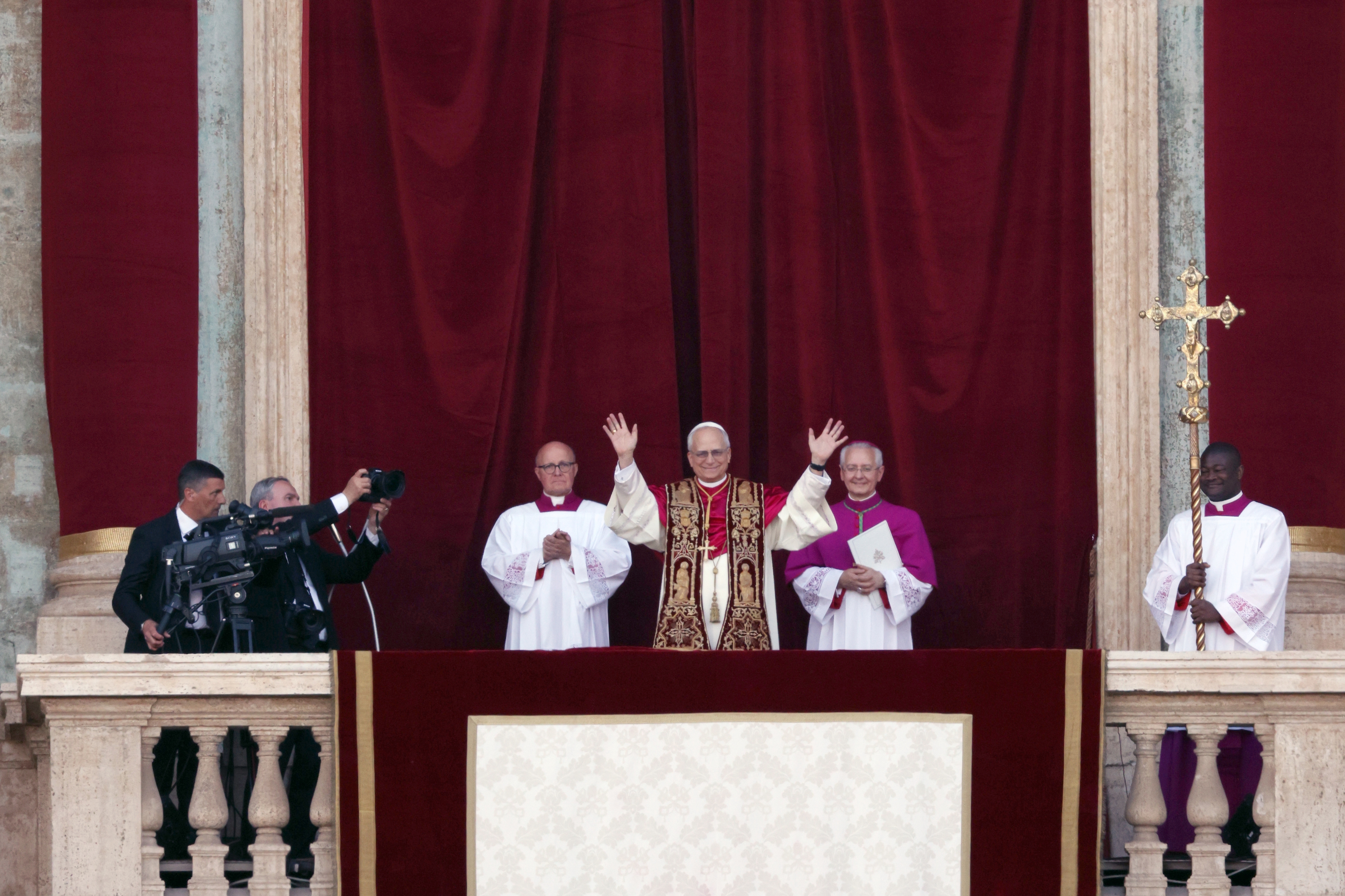 A religious leader in ornate vestments stands on a balcony, flanked by officials, raising hands to greet a crowd