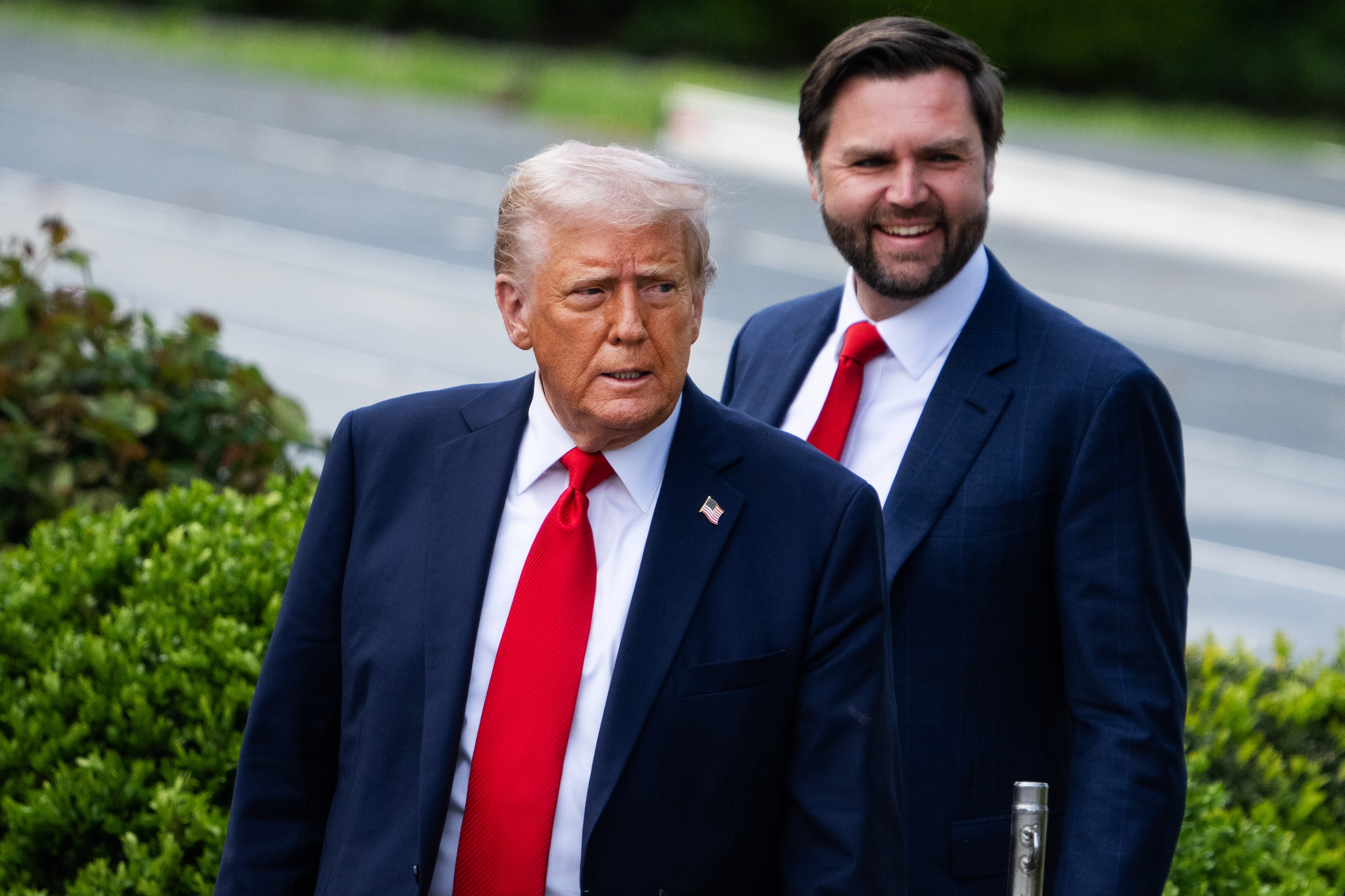 Two men in formal suits with red ties are walking outside. They appear engaged and attentive, suggesting a public or political event
