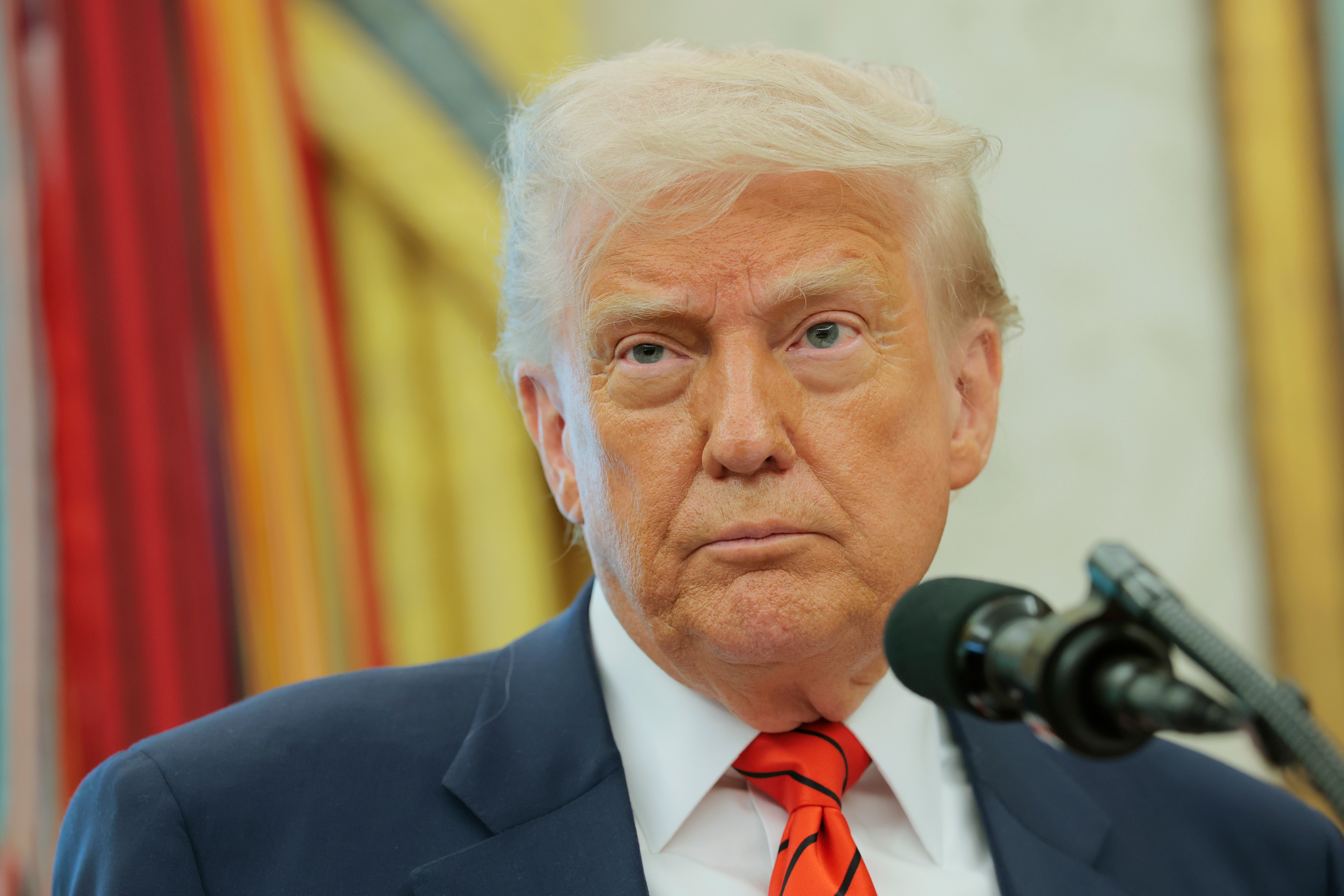 A man wearing a suit and striped tie stands at a microphone, appearing serious, in front of a decorated backdrop