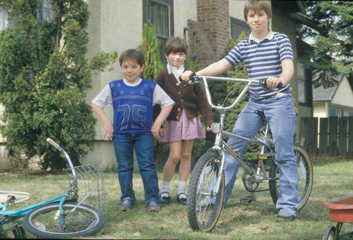 Three kids stand in a yard with two bicycles. One child wears a striped shirt and holds a bike, another wears a vest, and the third has a skirt and sweater