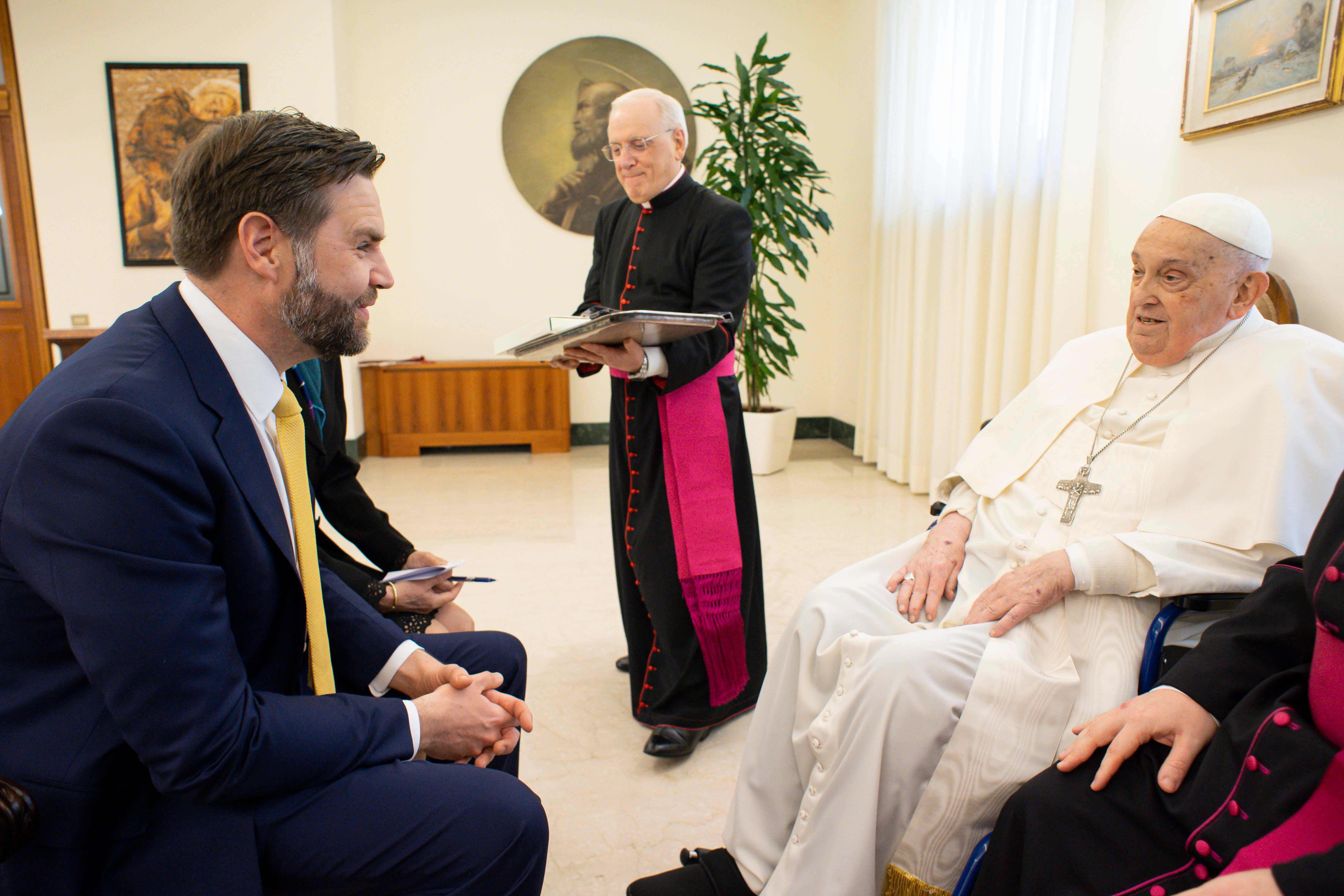 A seated religious leader in white attire converses with a man in a blue suit and yellow tie in a formal setting. A priest stands nearby holding a book