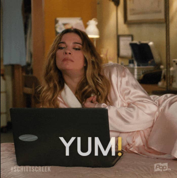 Person sits in a bed, wearing a silky robe, looking at a laptop. The word "YUM!" is displayed on the screen