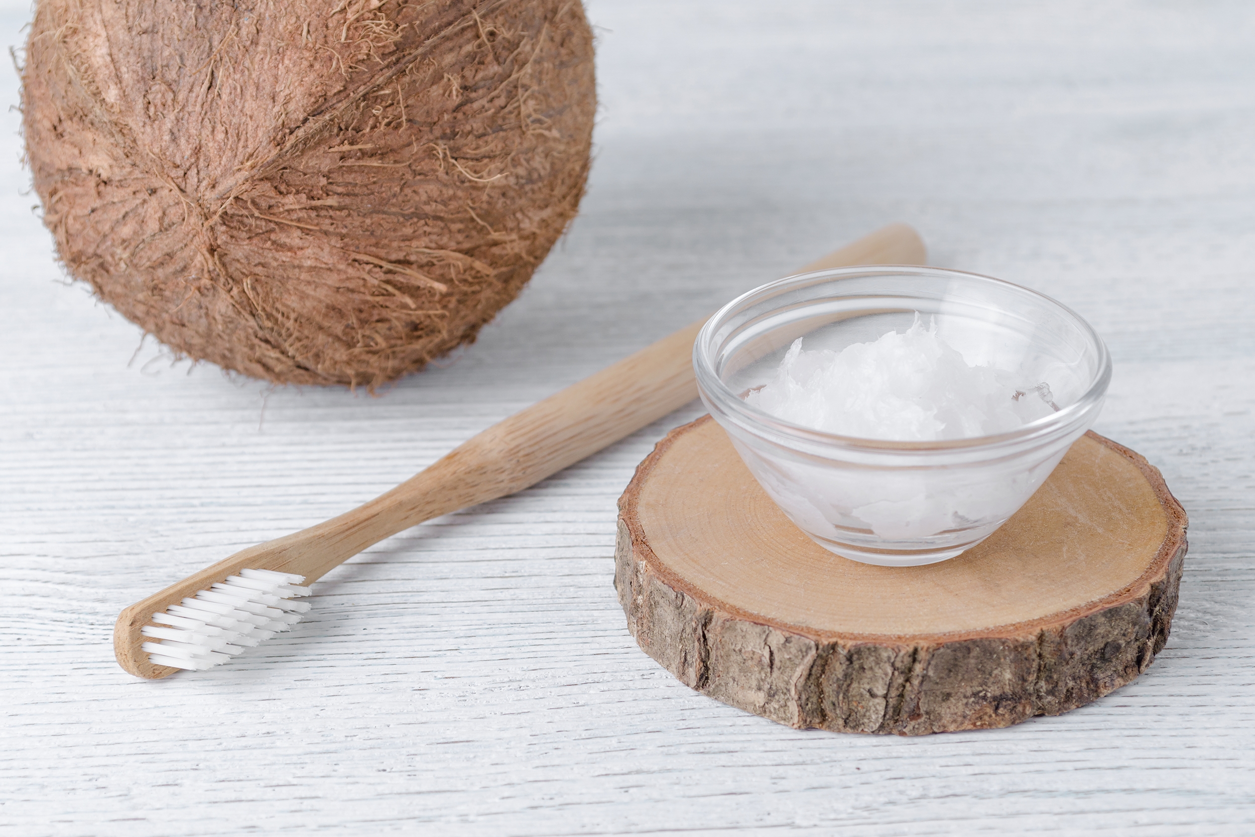 A toothbrush and a bowl of coconut oil are displayed on a wooden slab with a coconut in the background, suggesting natural dental care