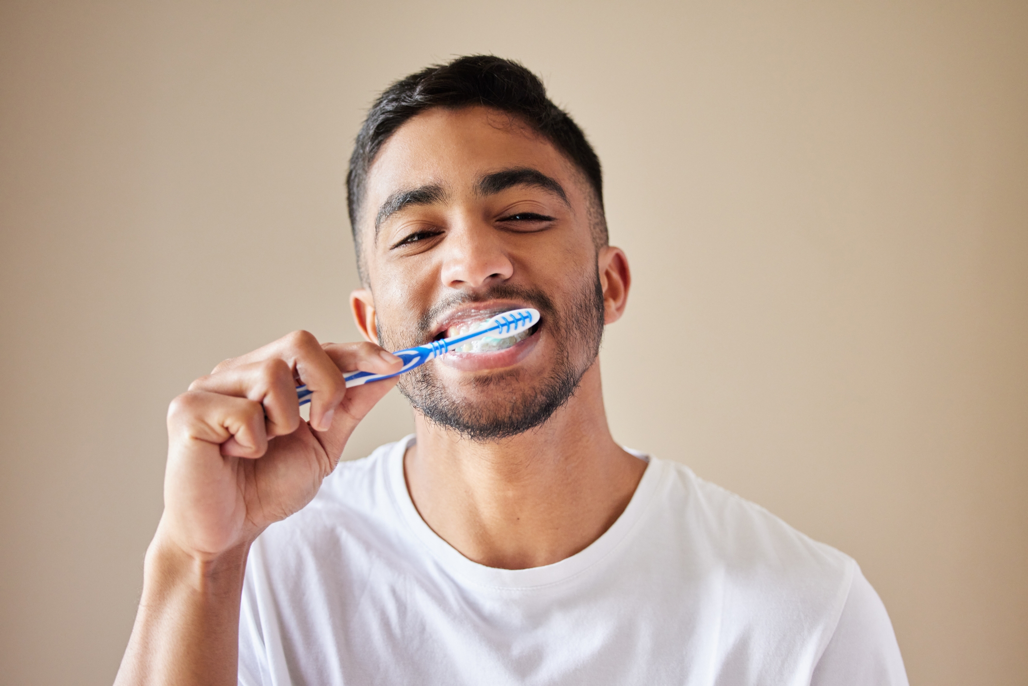 Person smiling while brushing their teeth with a blue toothbrush, wearing a plain white t-shirt