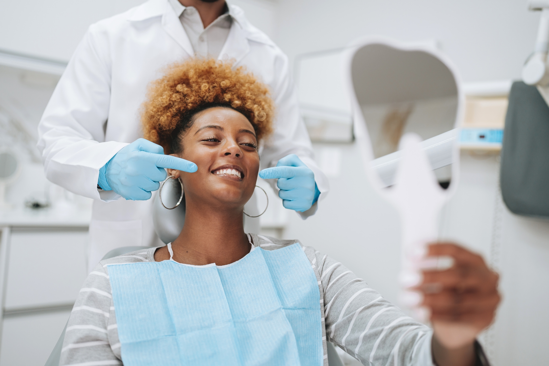 Person in a dentist's chair, holding a mirror, while a dentist points to their teeth, indicating satisfaction with the dental work