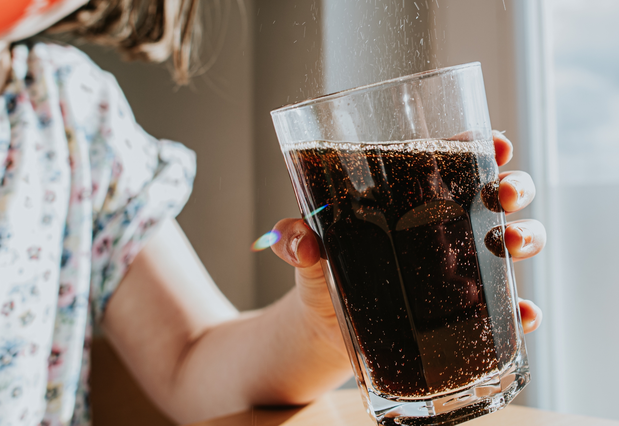 Person holding a glass of fizzy soda, capturing bubbles rising. Daylight filters through the window, creating a casual and refreshing scene