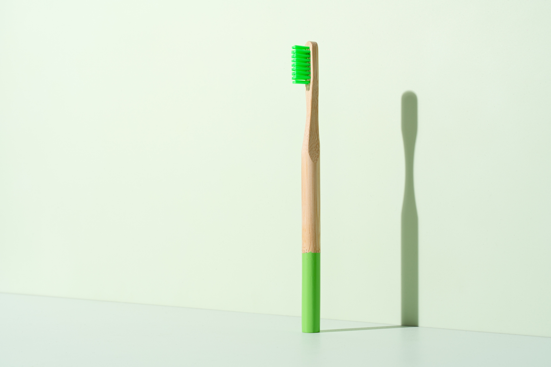 A bamboo toothbrush with a green bristle head and handle base stands upright on a light surface, casting a shadow