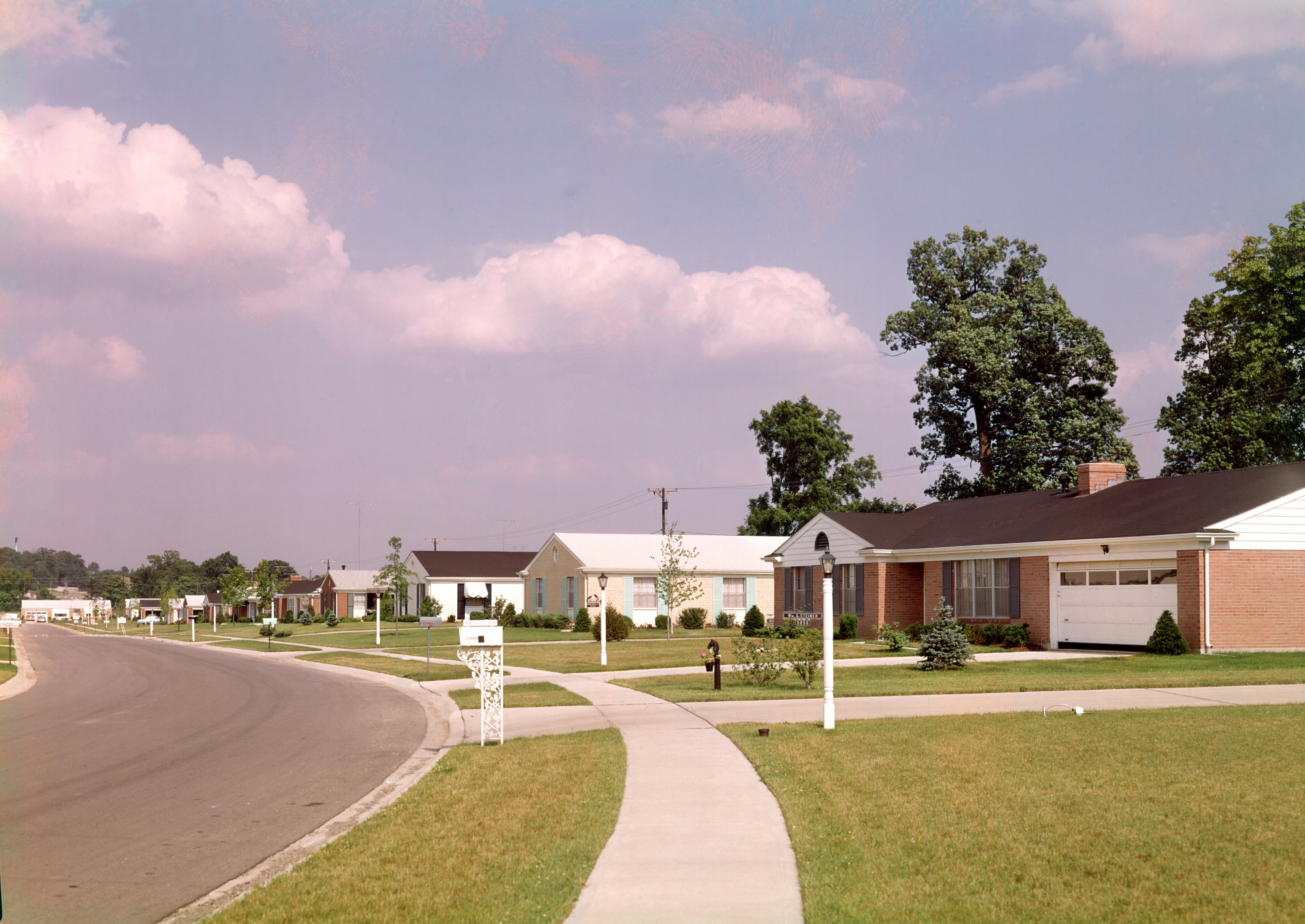 Suburban street with mid-century homes, large lawns, and trees lining the road under a partly cloudy sky