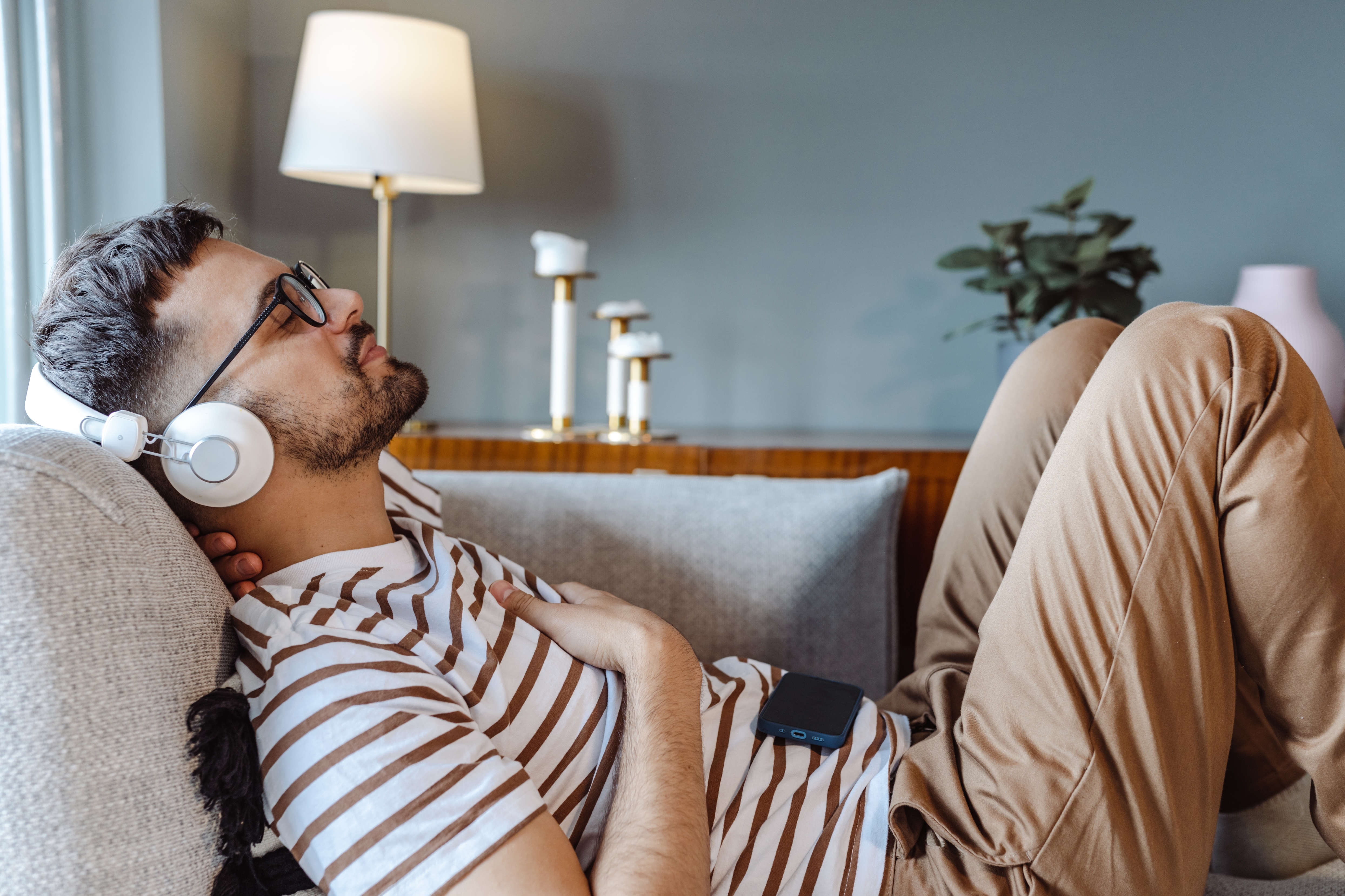 Man relaxing on a couch wearing headphones and a striped shirt, with eyes closed and hands on chest. A lamp and plants are in the background