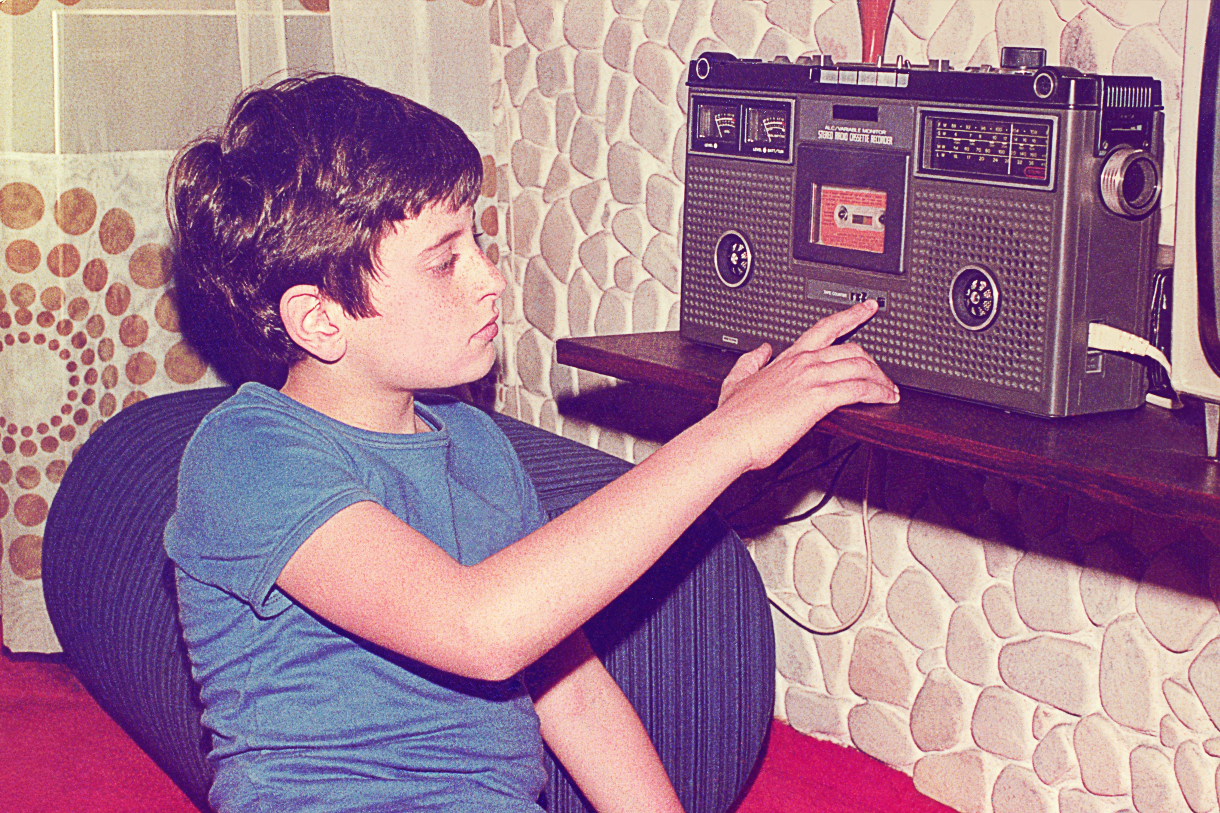 Boy listening to a tape on a retro cassette player in a room with patterned wallpaper