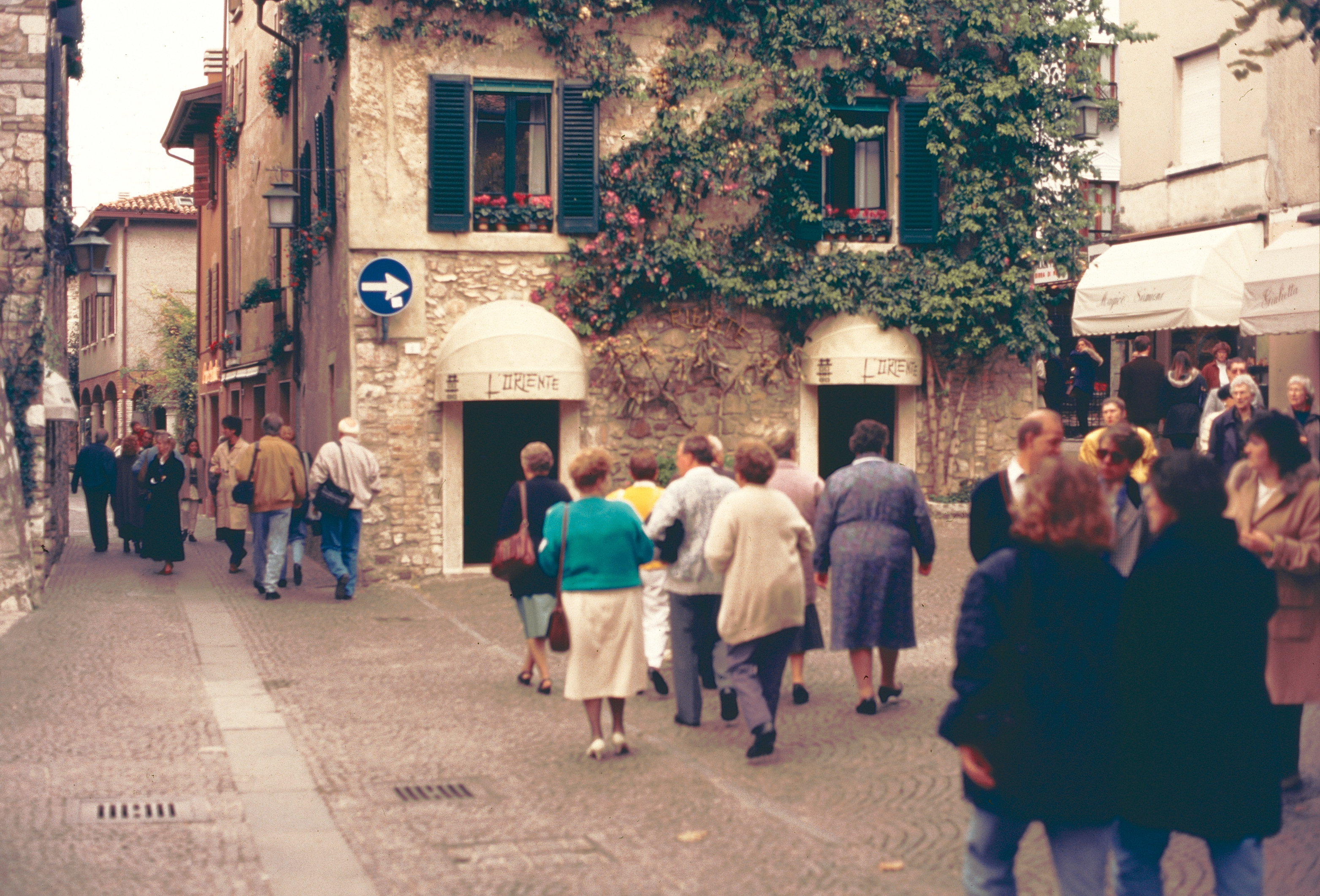 People walk along a quaint cobblestone street lined with stone buildings and greenery