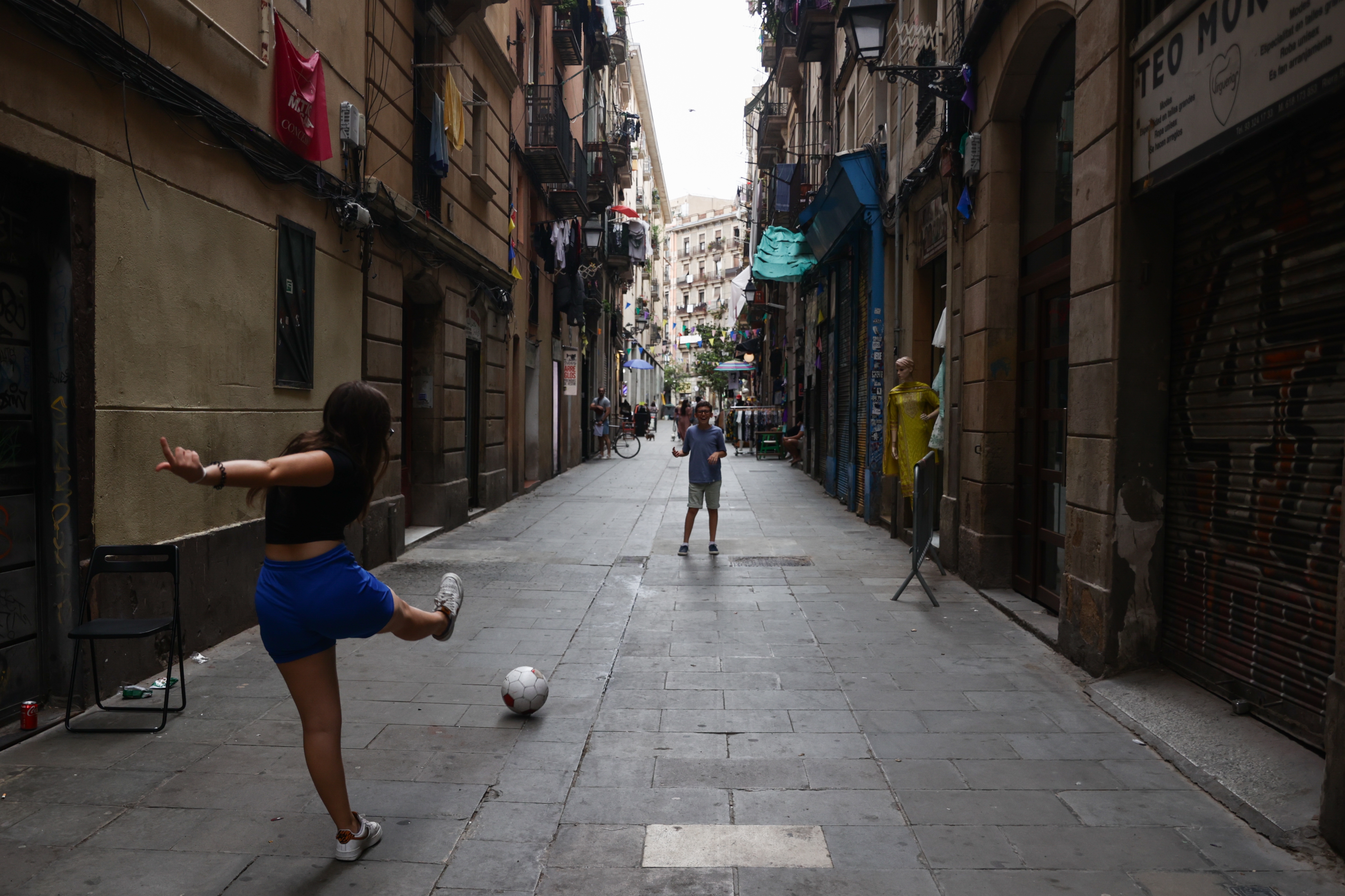 Person kicking a soccer ball to another in an urban alleyway lined with clothes on balconies, creating a lively and casual atmosphere