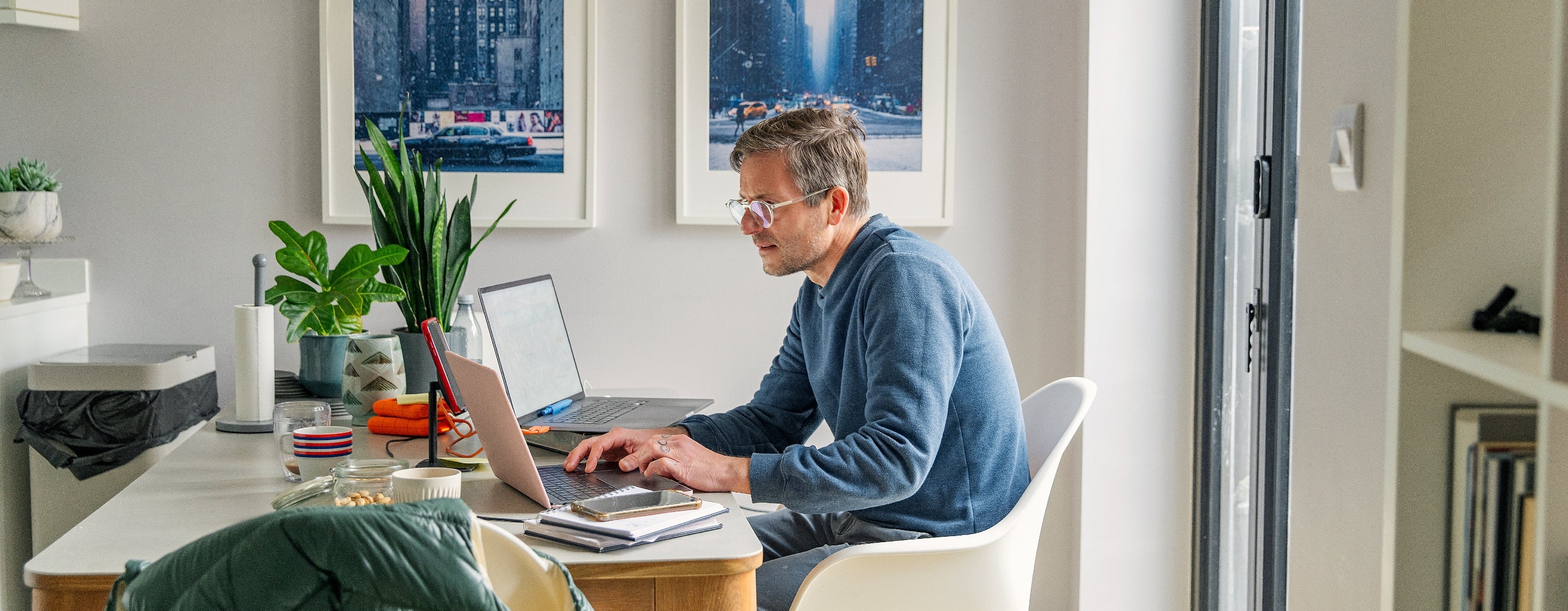 A person works on a laptop at a dining table in a modern, bright room with cityscape art and plants in the background