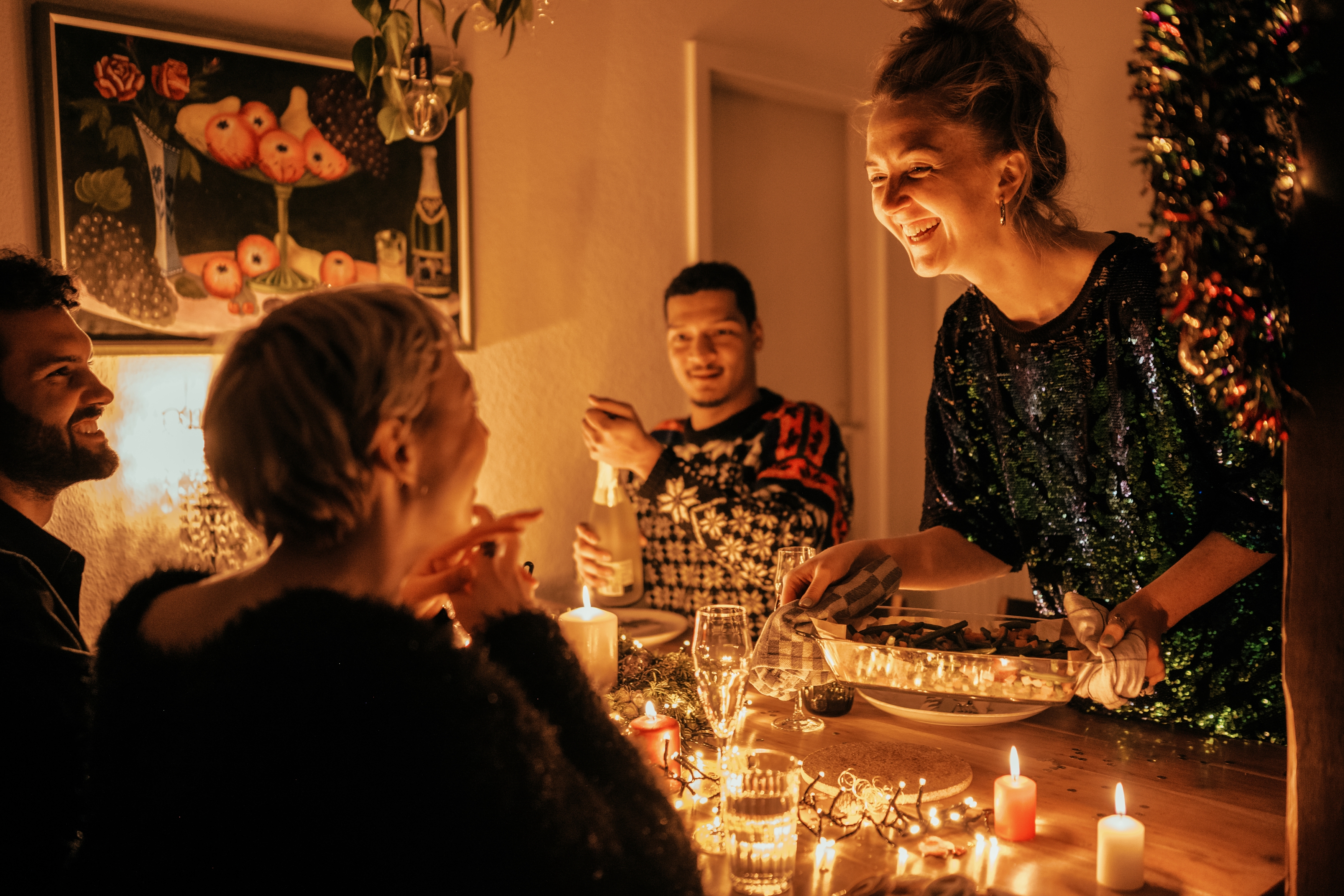People laughing and dining at a candlelit table, with one person serving food while others converse and enjoy the festive atmosphere