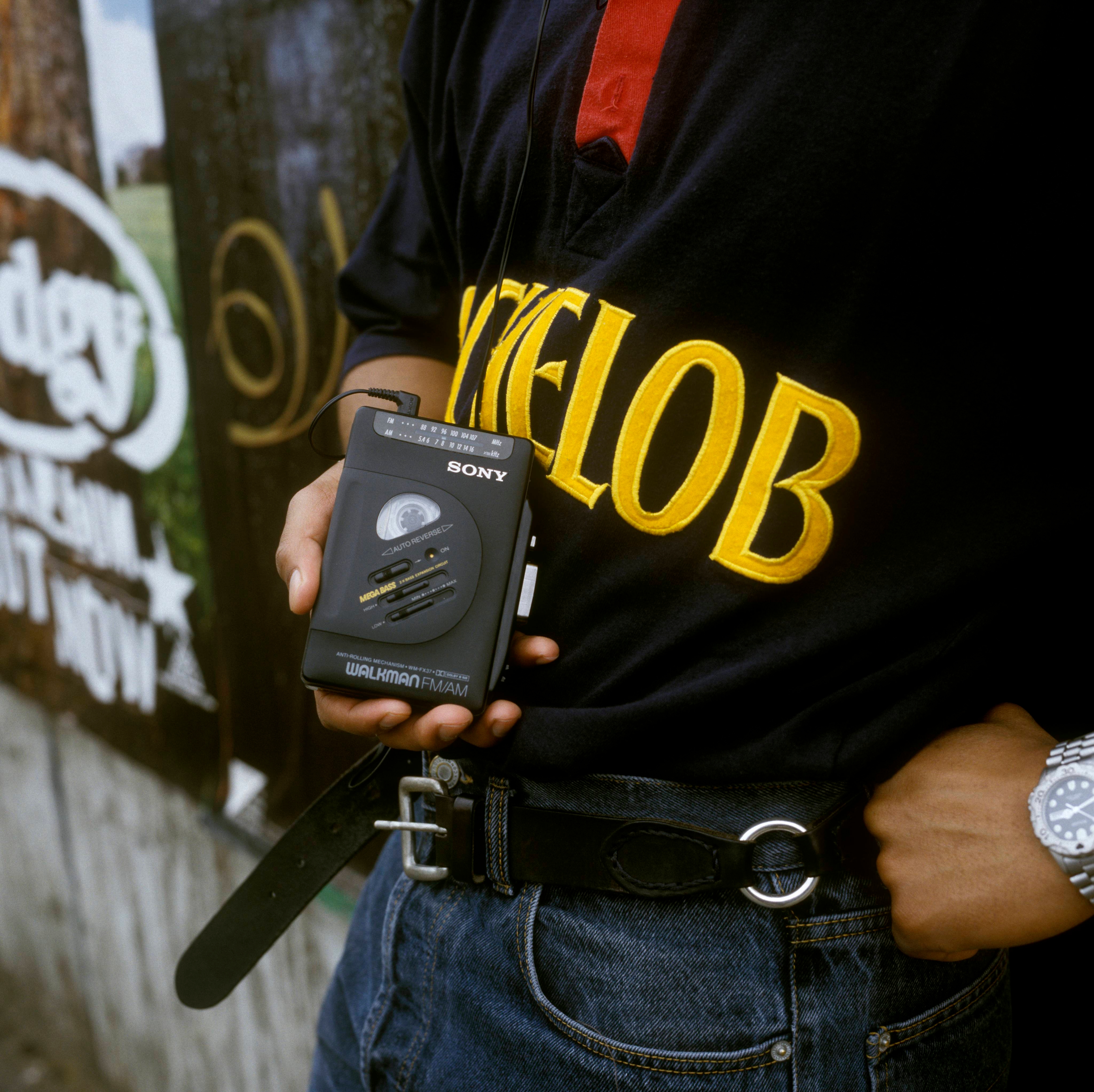 Person wearing a &quot;Nightclob&quot; shirt holds a Sony Walkman and wears a wristwatch and jeans with a belt