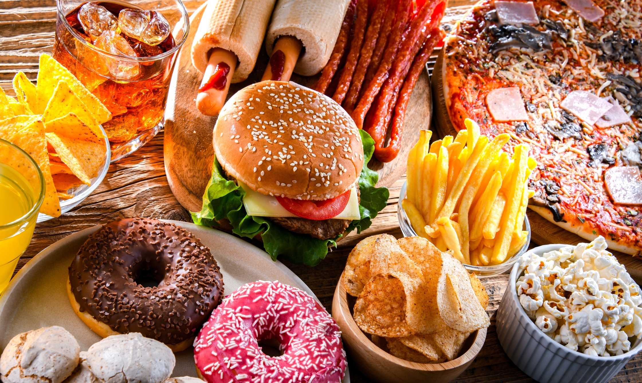 Assorted junk food on a table, including burgers, pizza, fries, hot dogs, donuts, popcorn, chips, and drinks, suggesting a laid-back travel snack theme