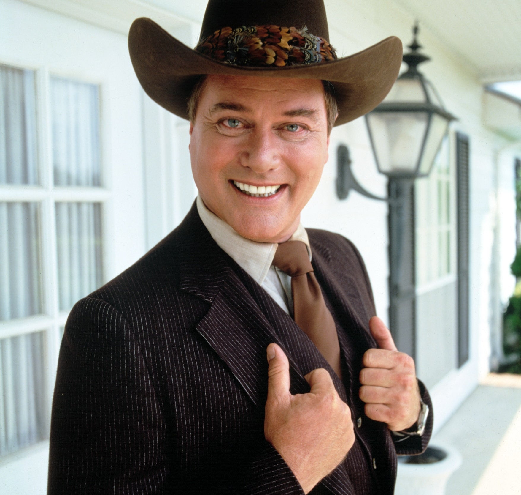 Person smiling in a pinstriped suit and cowboy hat, standing on a porch with a lantern in the background