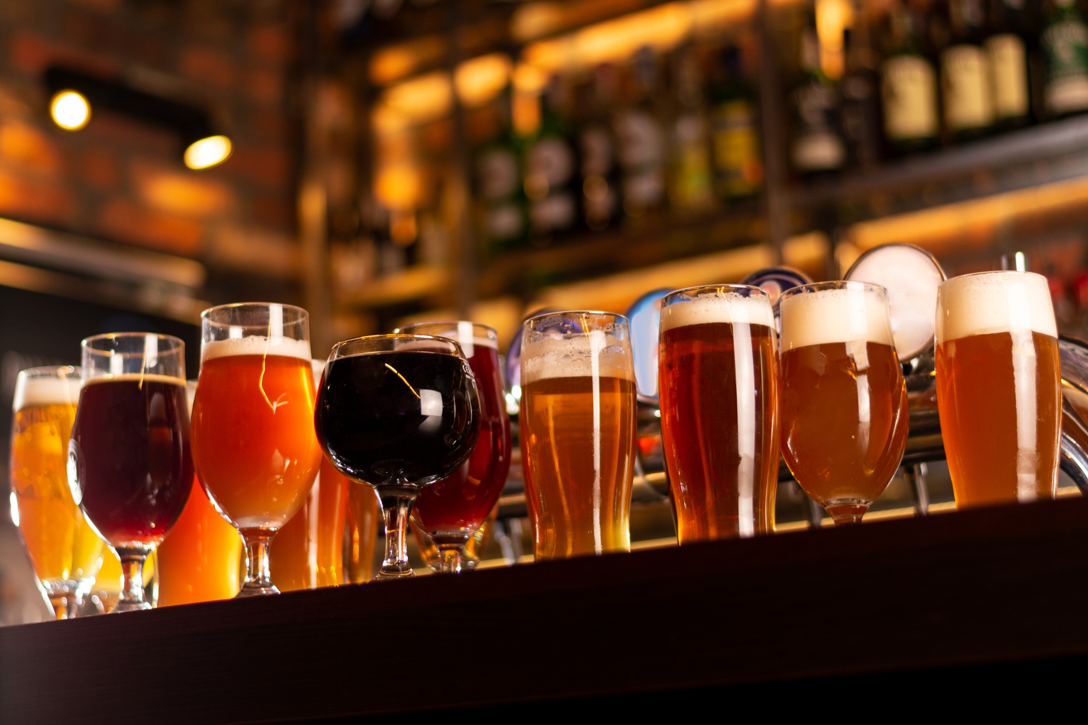 A row of various glasses filled with different types of beer on a bar counter, with a blurred background of shelves and lights in a bar setting