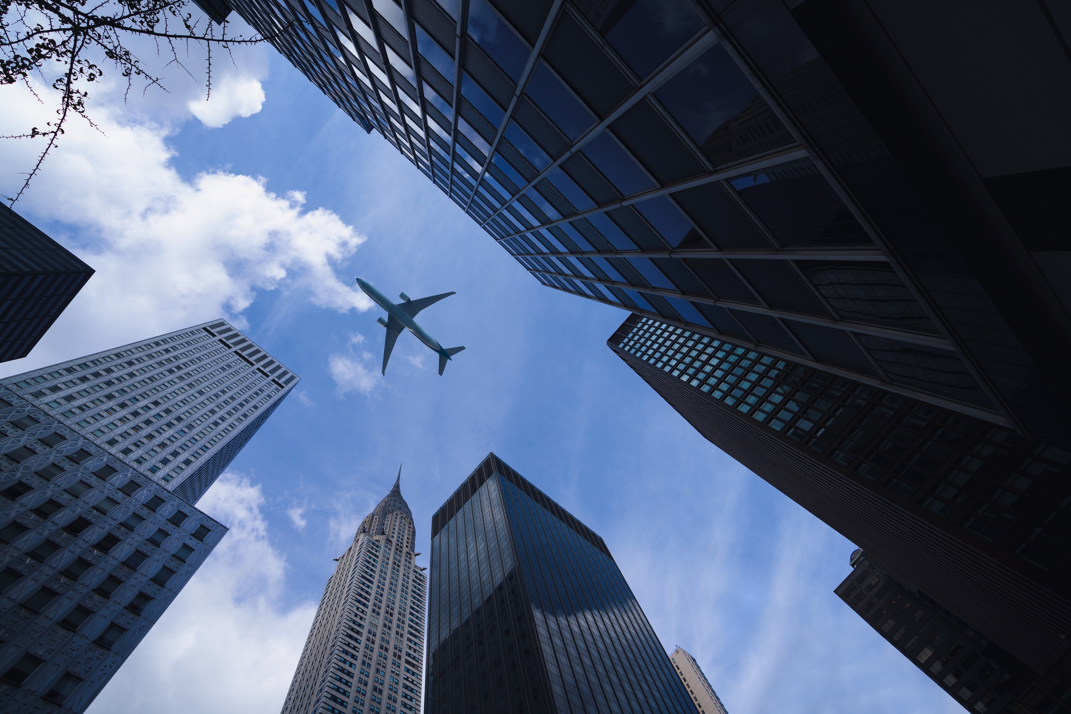 Airplane flying over tall skyscrapers, viewed from below, against a backdrop of a clear sky, capturing a travel theme