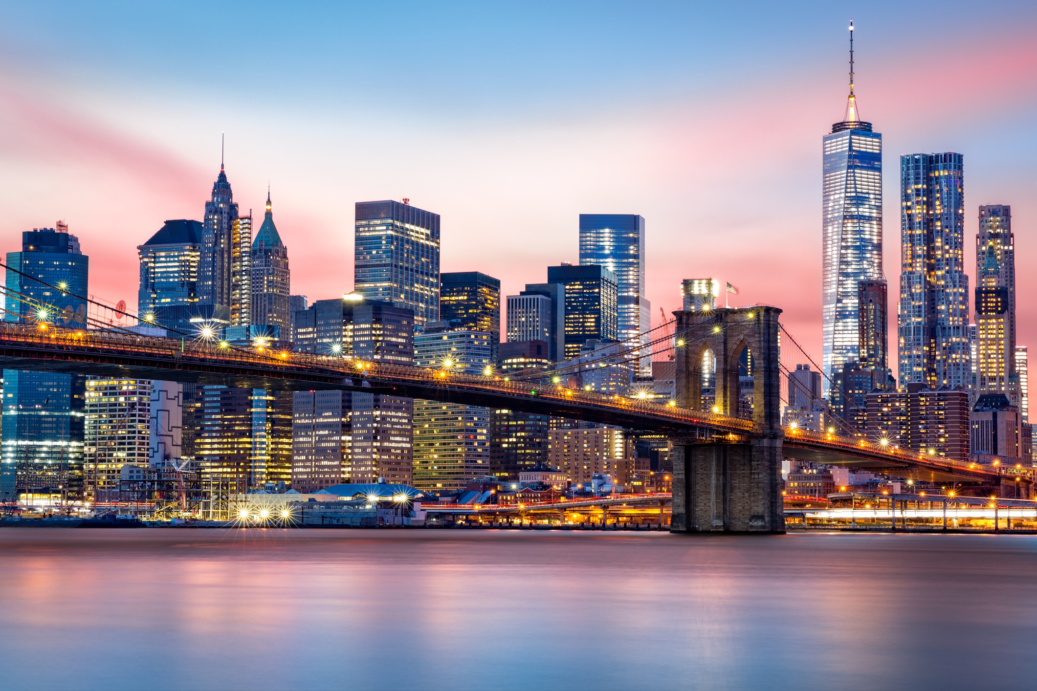 New York City skyline at dusk with illuminated skyscrapers and Brooklyn Bridge over the calm East River