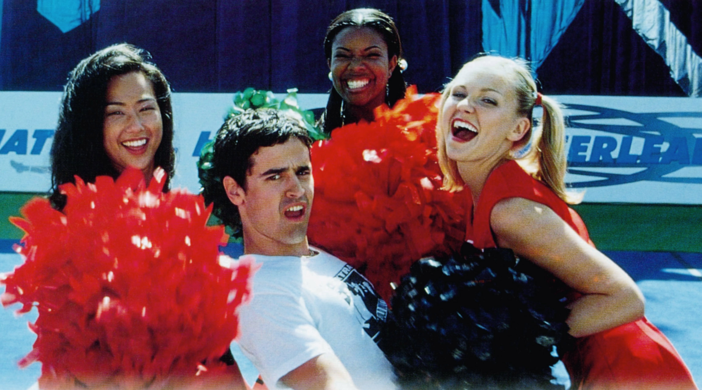 A group of cheerleaders surrounds a man in a fun pose, smiling and holding pom-poms, with a sporty background suggesting a lively event