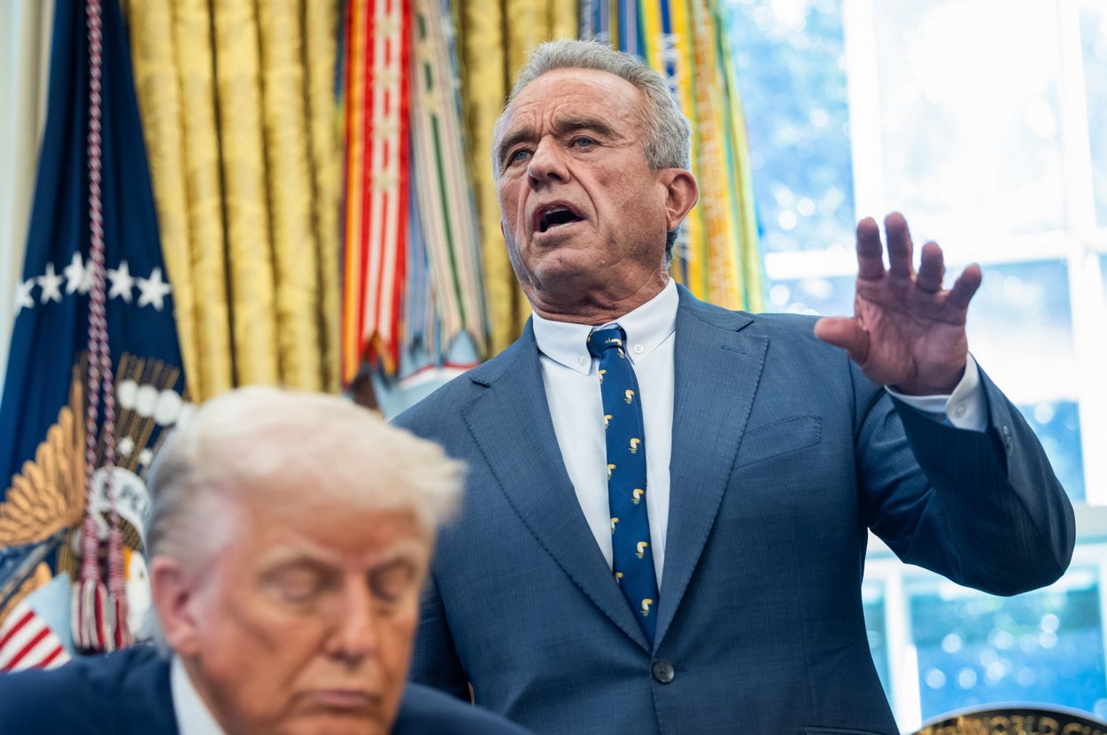 Two men in a formal setting, Robert Kennedy Jr. speaking animatedly in a suit, while President Trump is seated. Background features U.S. presidential decor.
