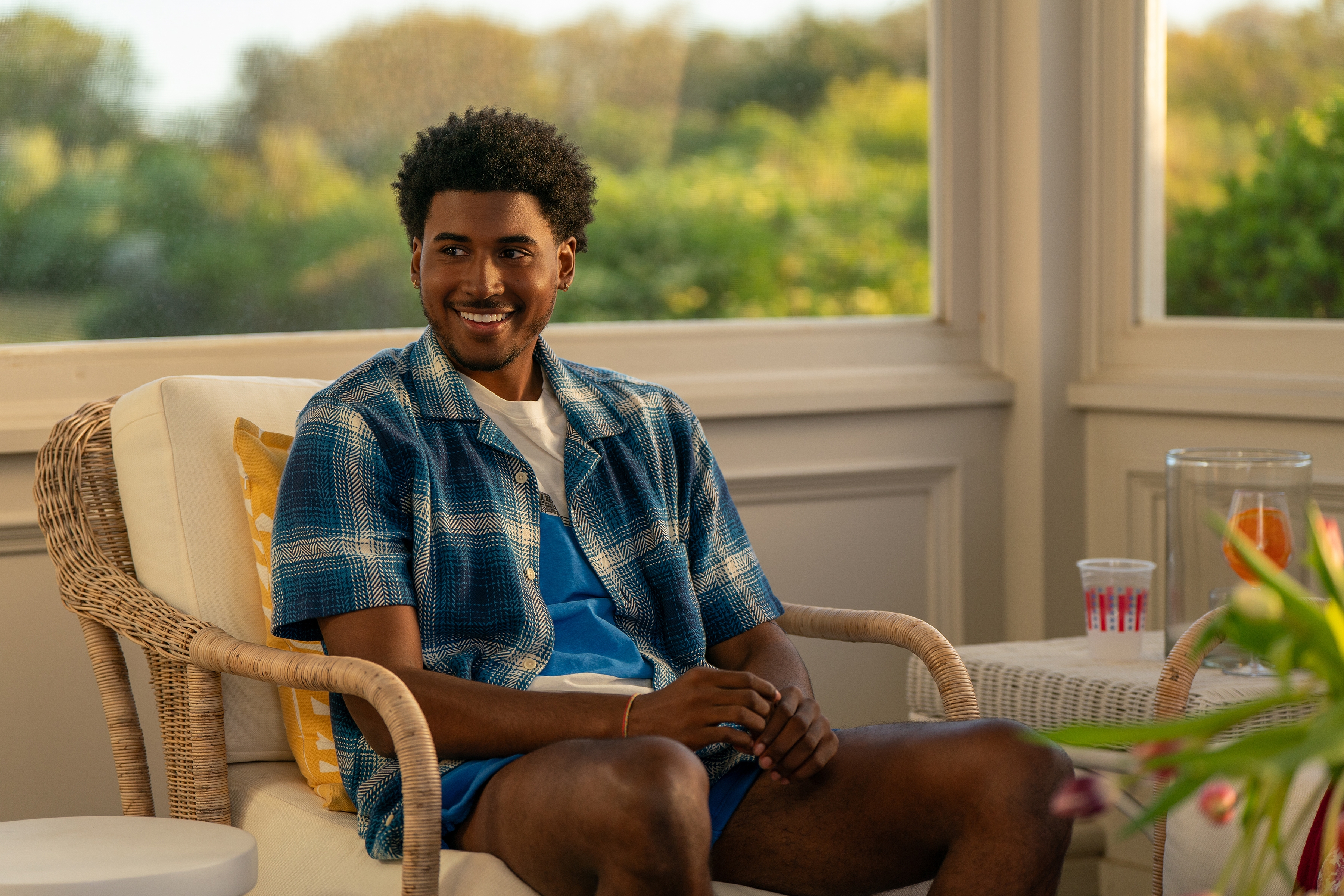 A person sits smiling in a wicker chair, wearing a casual checkered shirt over a blue outfit, in a cozy, sunlit room