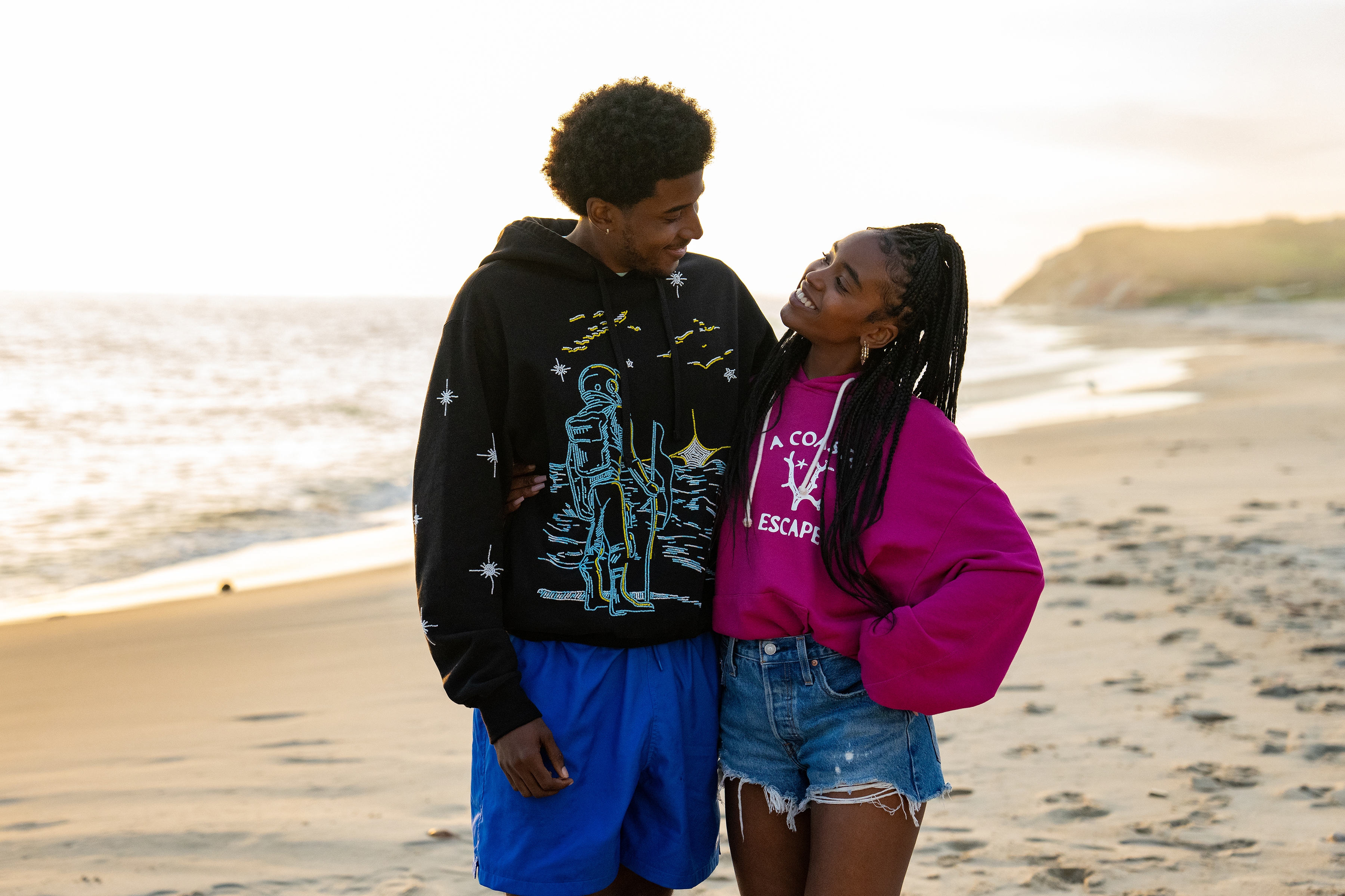 A couple stands on a beach, smiling at each other. One wears an astronaut-themed hoodie, the other a pink shirt with shorts