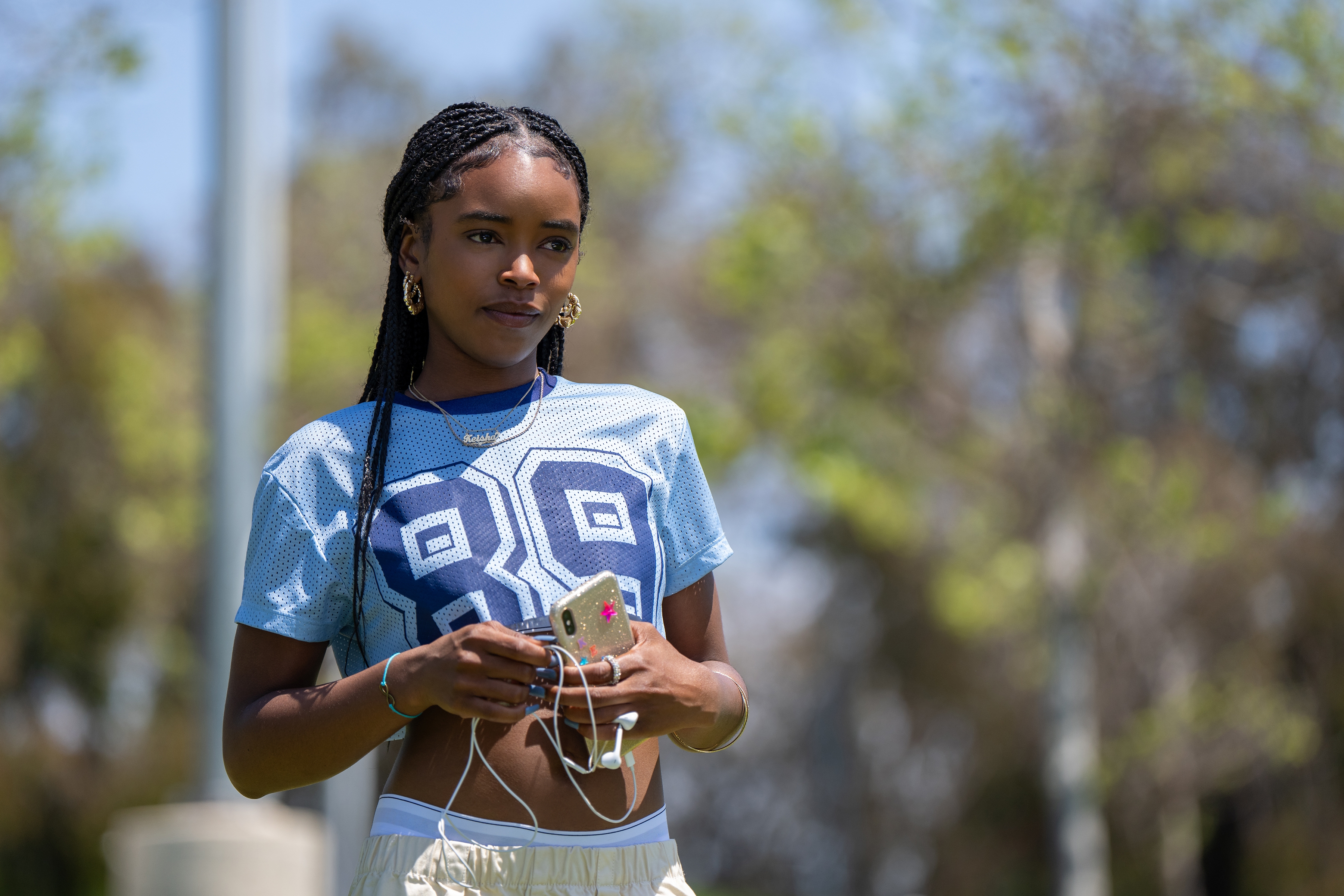 Person outdoors wearing a sporty top and holding a phone with earphones, looking thoughtful