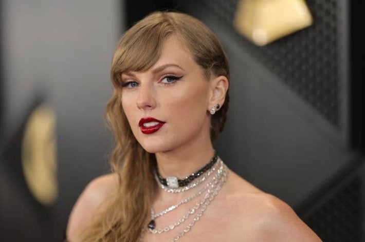 A person on a red carpet wearing a strapless outfit and layered necklaces, with styled hair and bold makeup