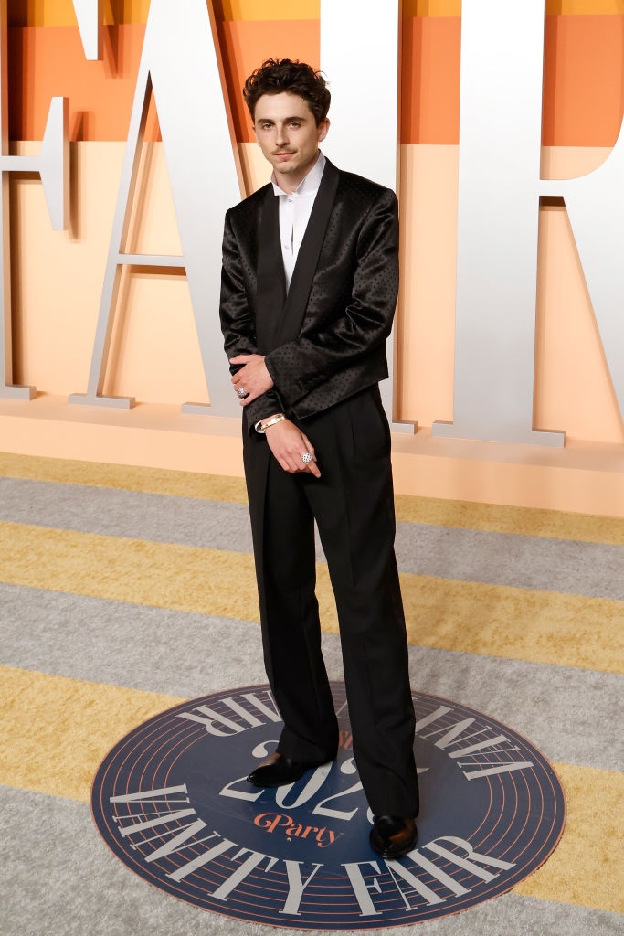 A person in a sleek tuxedo poses on the red carpet at an event, standing confidently with a neutral backdrop displaying large letters