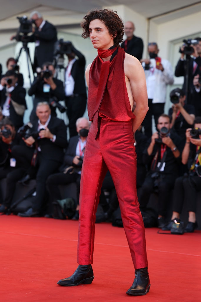 Person on red carpet in sleeveless, fashion-forward outfit with tailored pants and neck fabric detail; photographers in background