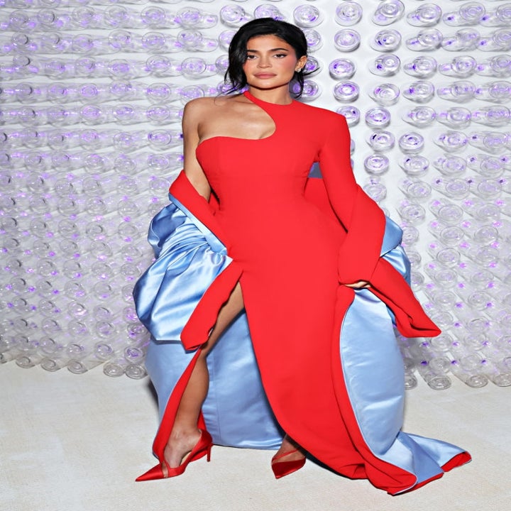 Person in a stylish asymmetrical red gown with blue train and side slit, posing in front of a wall with circular patterns