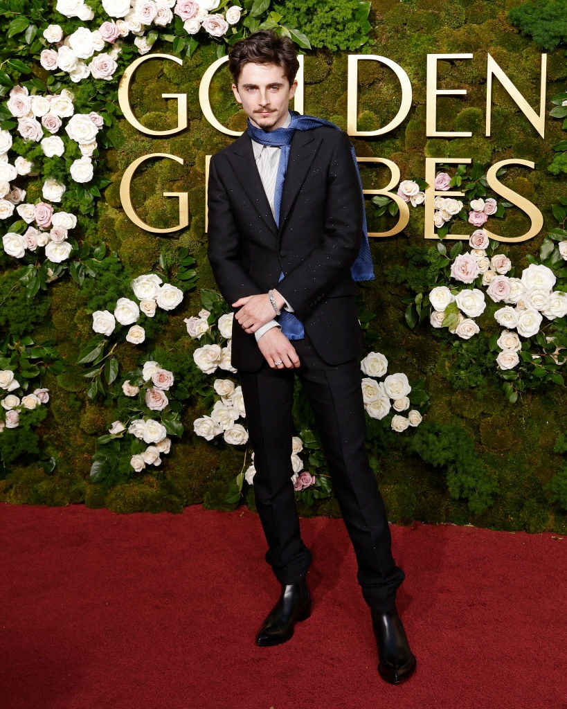 Person in a tailored suit with a blue scarf, posing on the red carpet at a floral backdrop event