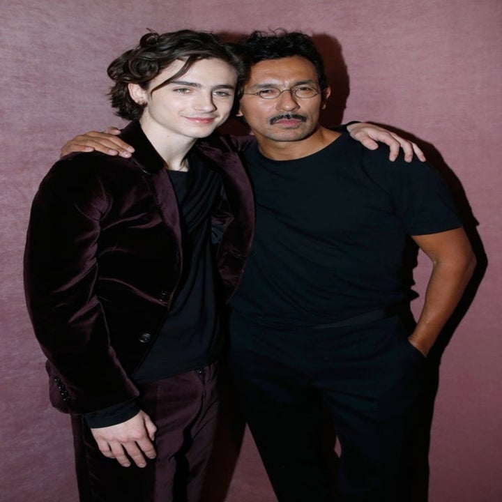Two individuals pose for a photo. One wears a velvet suit, and the other is in a casual outfit