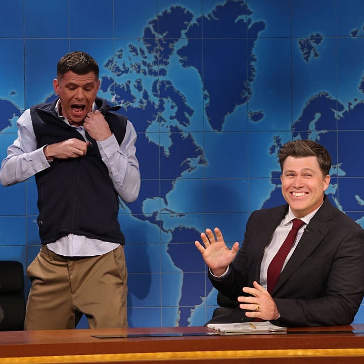 Two men on a comedy show set, one in a suit laughing and the other playfully adjusting his sweater vest. World map backdrop