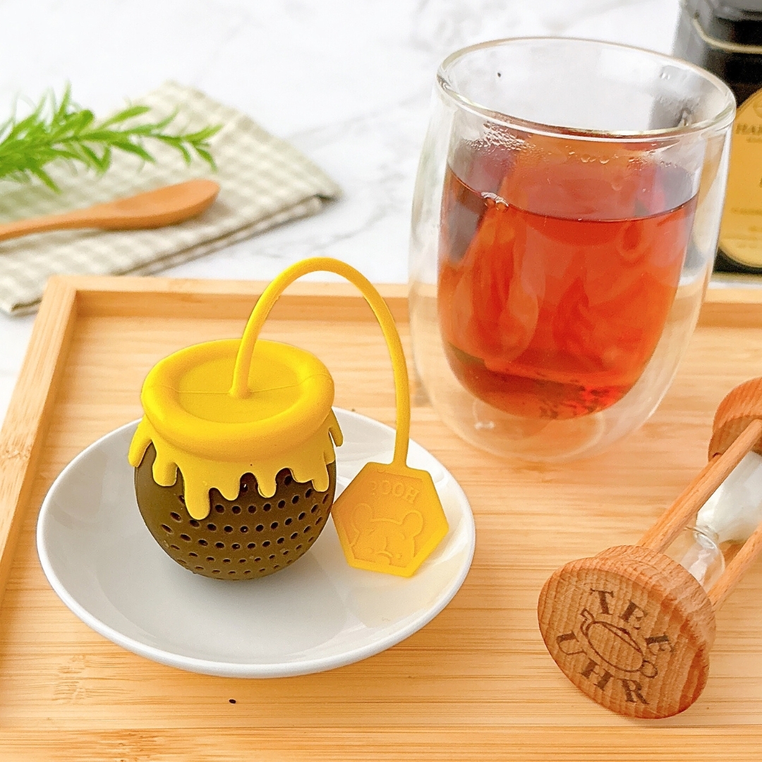 はちみつ壺の形をしたティーインフューザーが皿に置かれ、隣に紅茶の入ったガラスカップと砂時計がある。