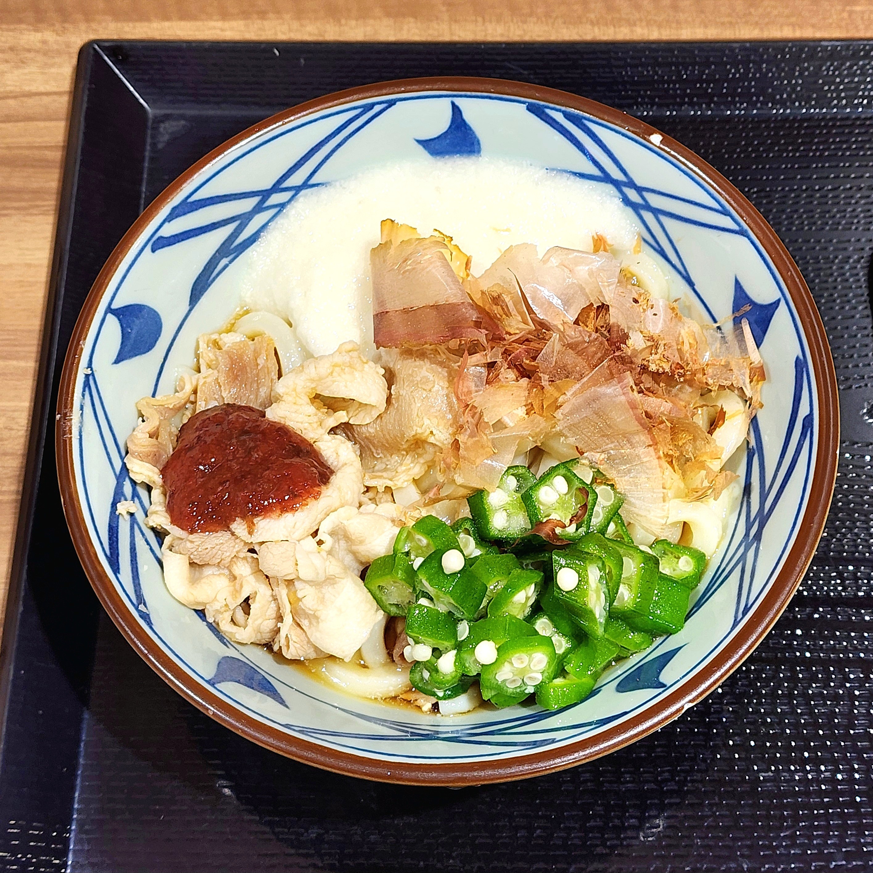 とろろうどんが盛られた青い模様の器に、具材として豚肉、オクラ、鰹節が添えられている様子。