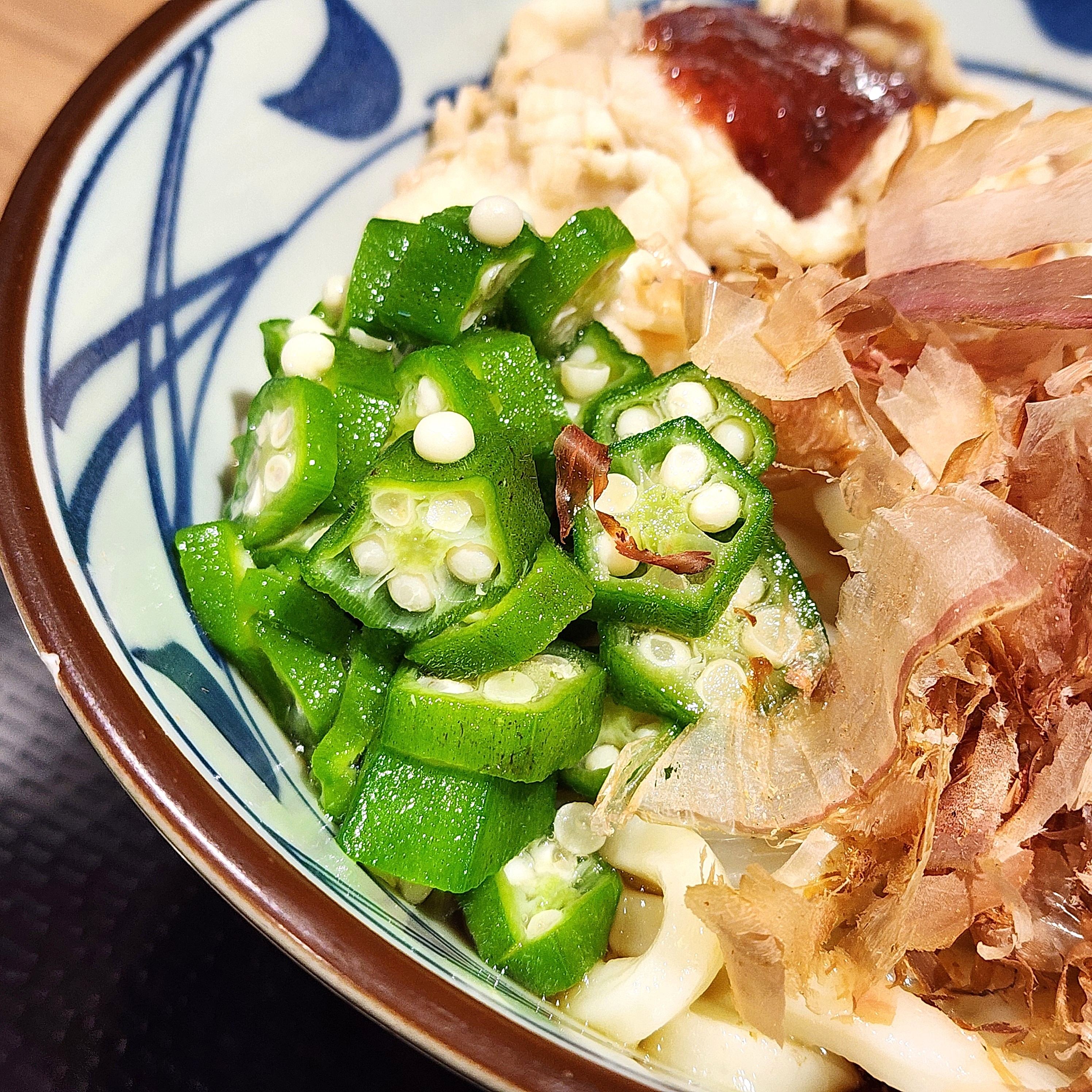 うどんの上にオクラ、鰹節、ソースがかかった具材が盛られている。