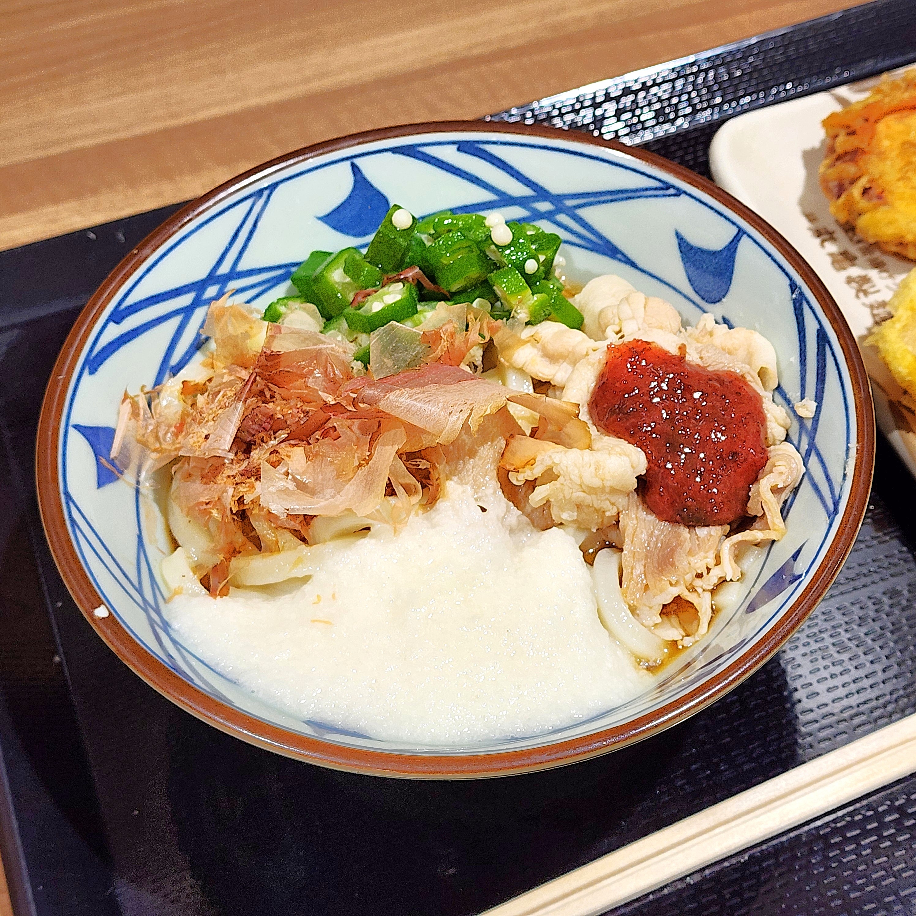 うどんの上に豚肉、山芋、オクラ、かつお節、赤いソースがのった料理。隣に揚げ物もある。